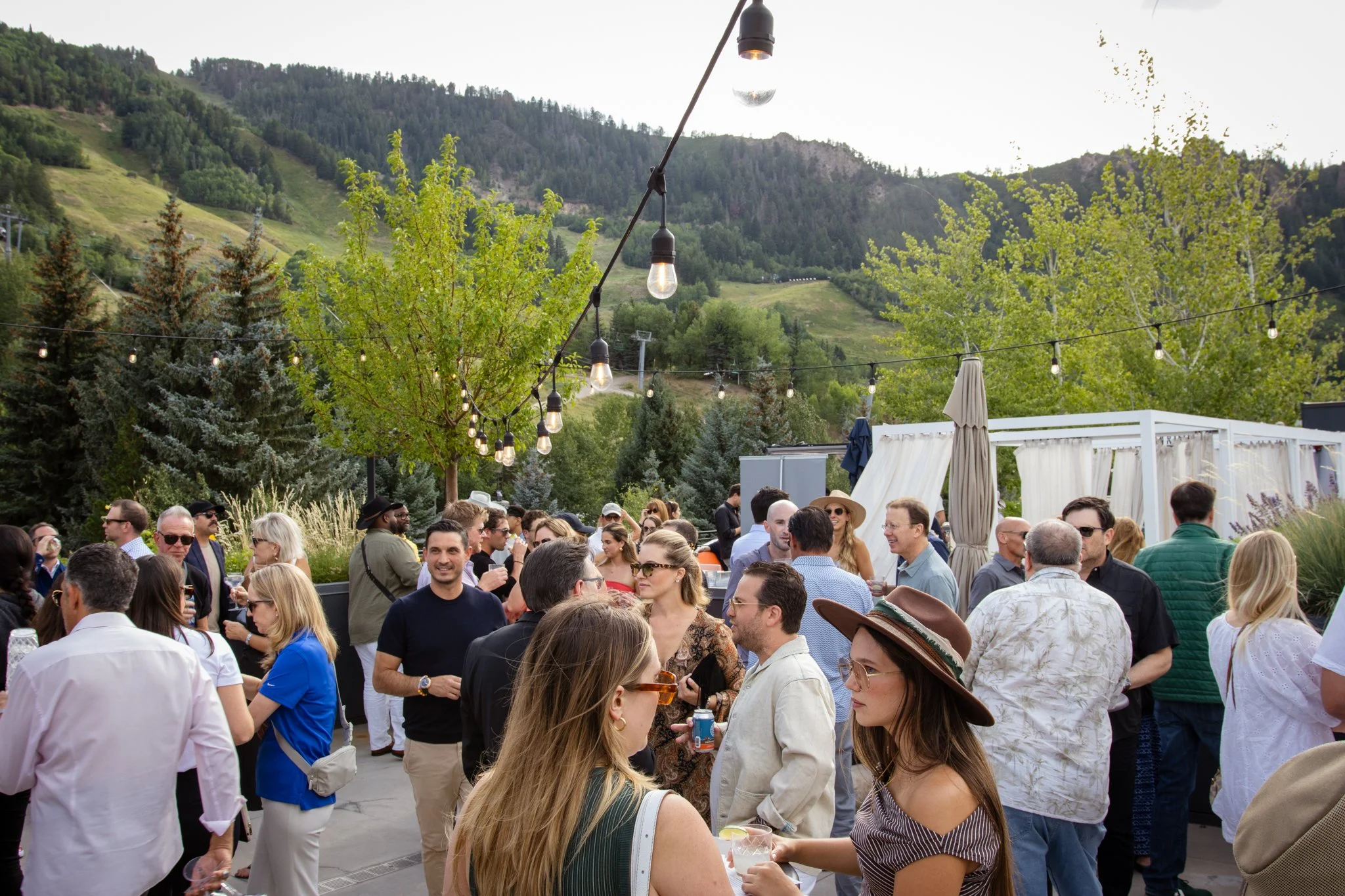 Outdoor gathering of people at a social event on a patio with string lights, trees, and mountains in the background during daytime