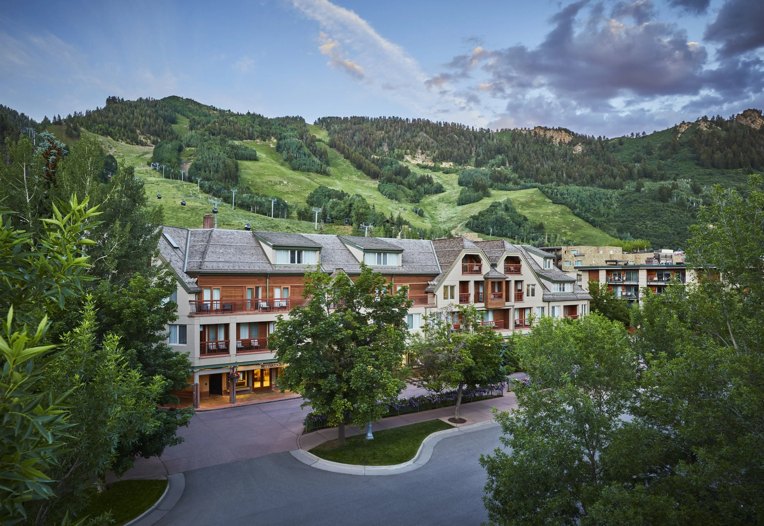 A mountain village with colorful houses surrounded by greenery and trees, with ski slopes and chairlifts in the background under a partly cloudy sky.