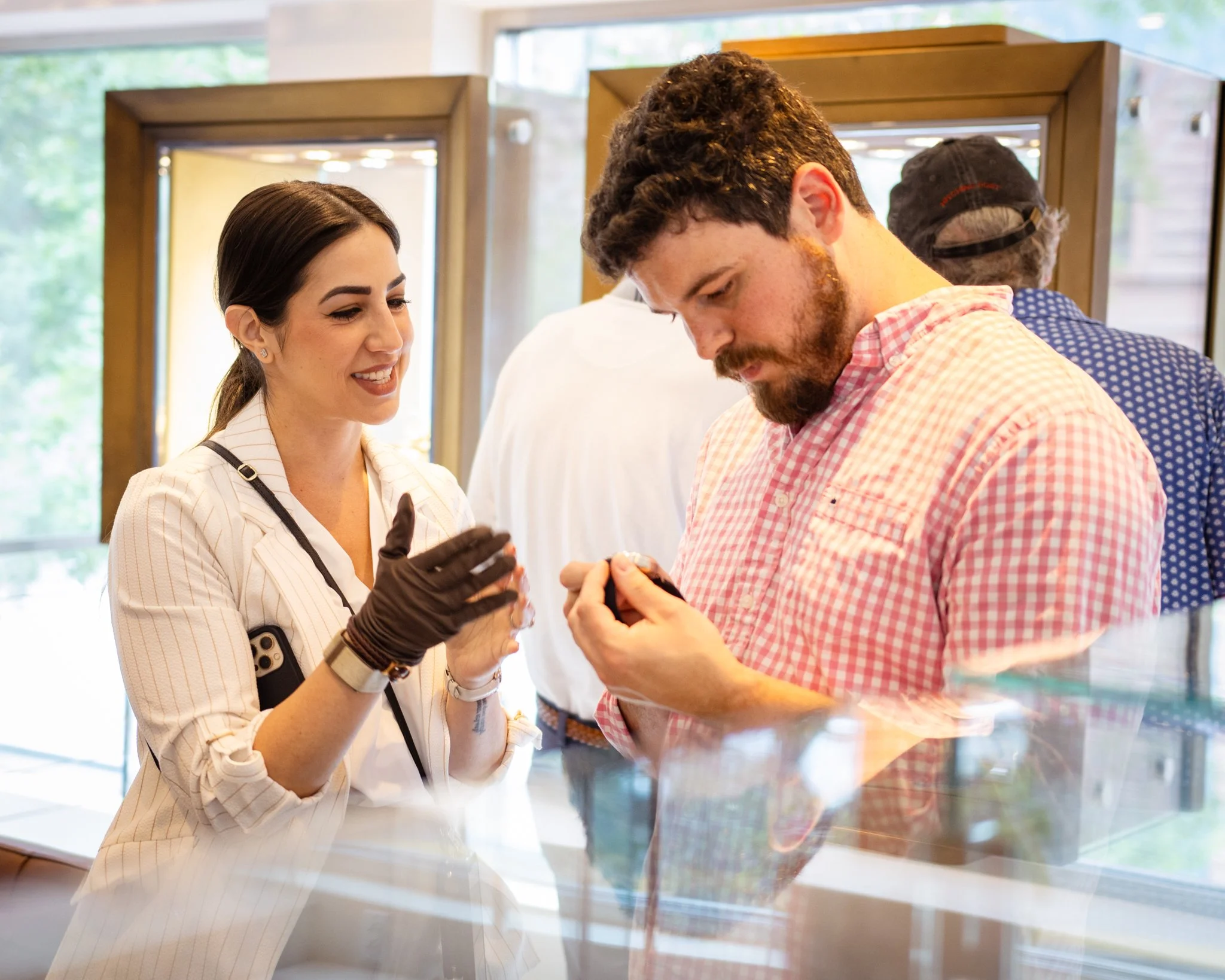 A woman with dark hair in a low ponytail, wearing a beige striped blazer and black gloves, smiling as she talks to a man with curly brown hair and a beard, wearing a pink checkered shirt, looking at a small item in his hands. They are inside a well-lit shop with large windows, other people in the background, and glass display cases.
