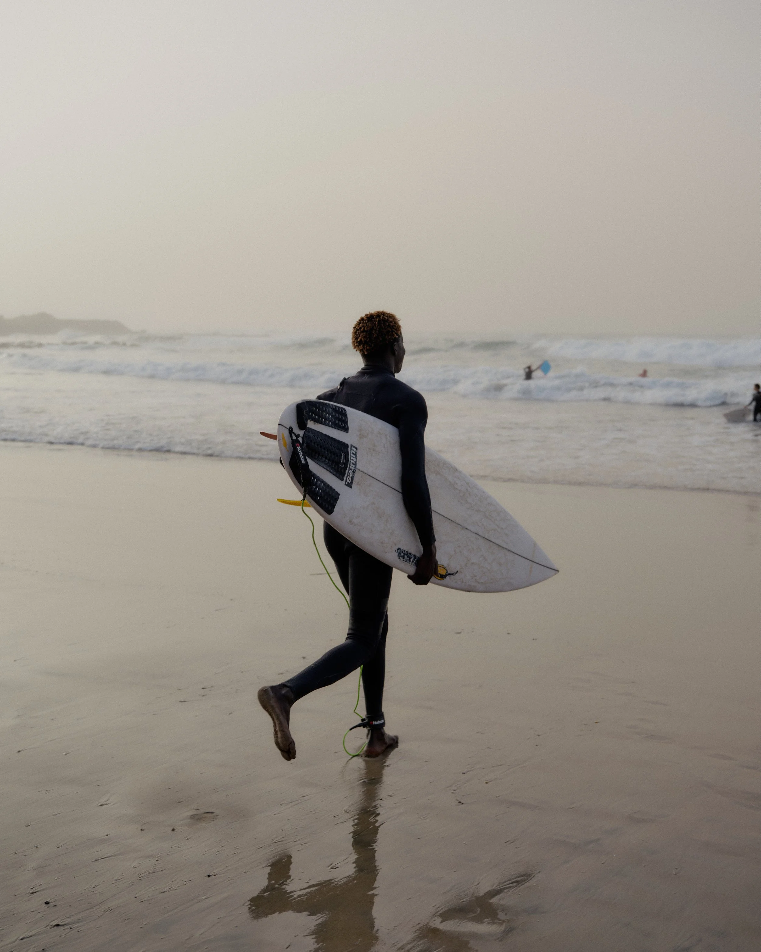 A person in a wetsuit carrying a surfboard walking along the beach toward the ocean, with other surfers in the background.