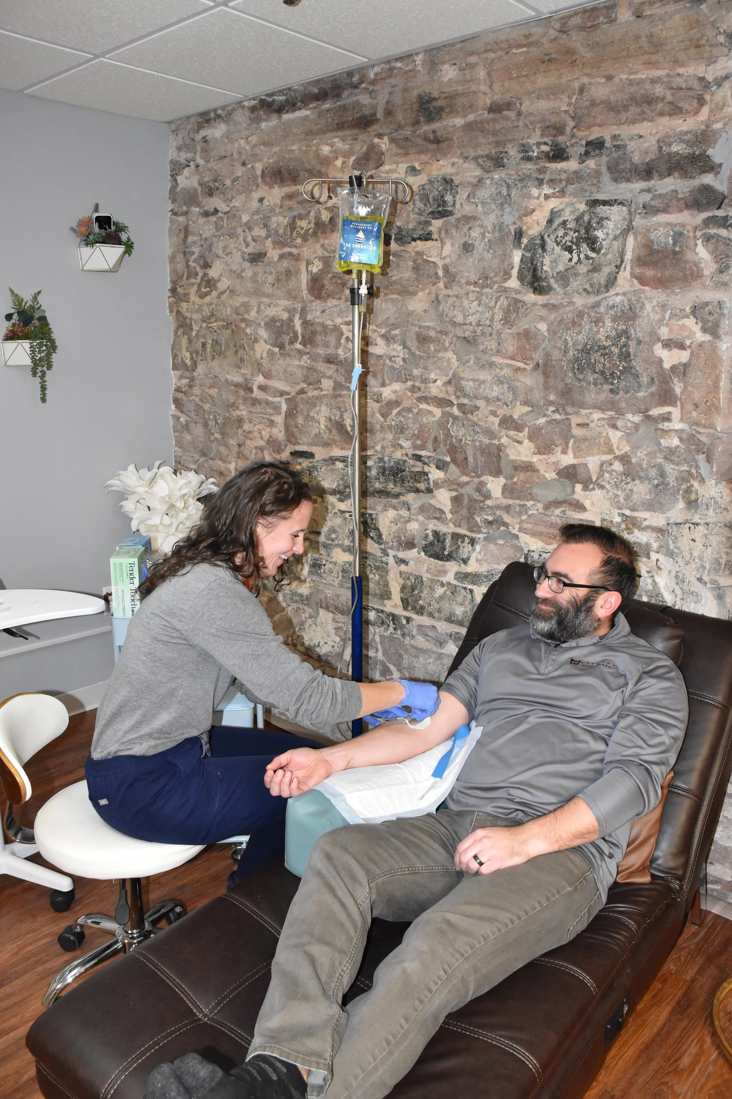 A woman is administering an IV to a man who is sitting on a reclining medical chair. The man has a bandage on his arm and looks relaxed. There is an IV stand nearby, and the setting appears to be a medical clinic with a brick wall background.