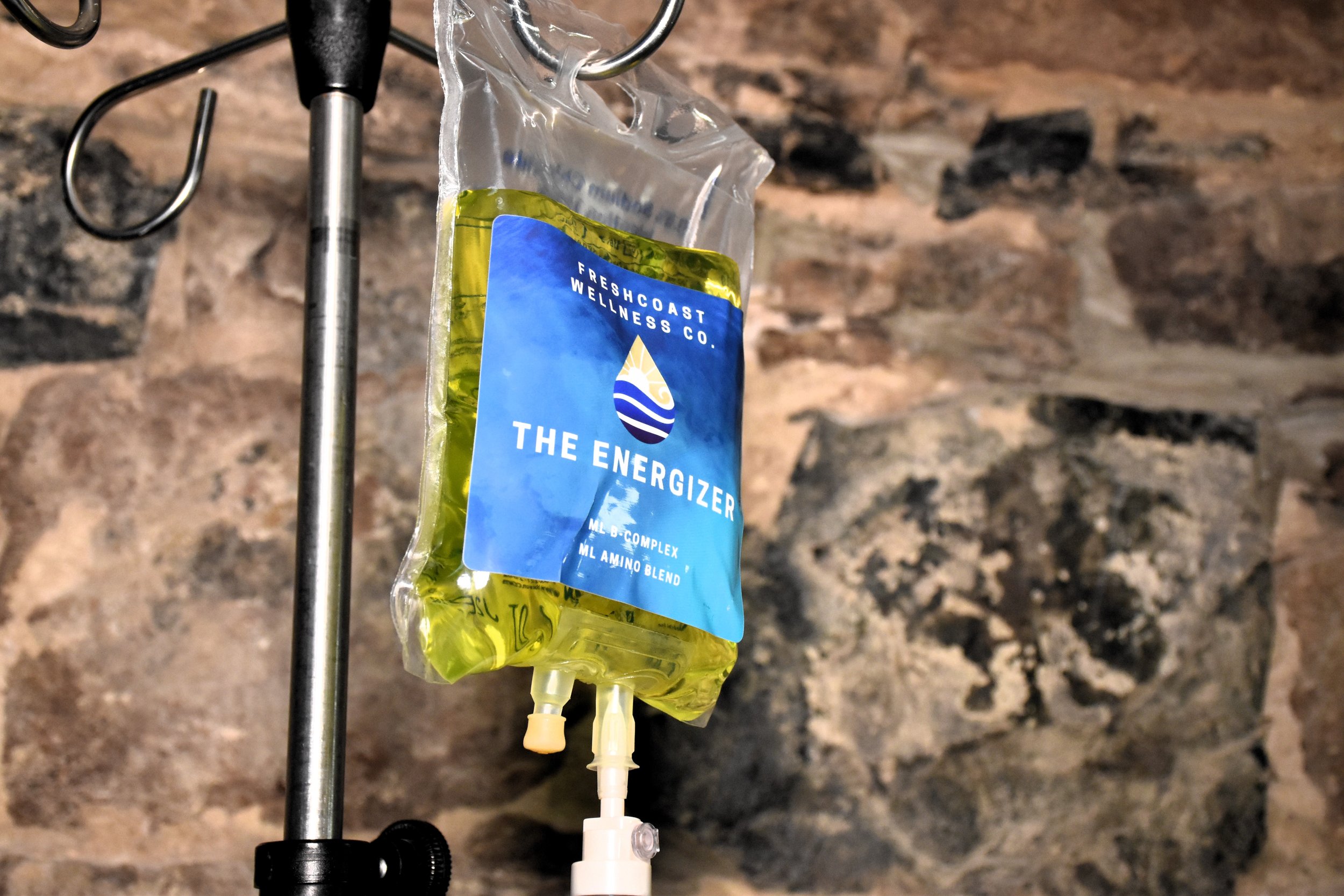 A hydration bag labeled 'The Energizer' hanging from a metal hook, filled with yellow liquid, against a stone wall background.