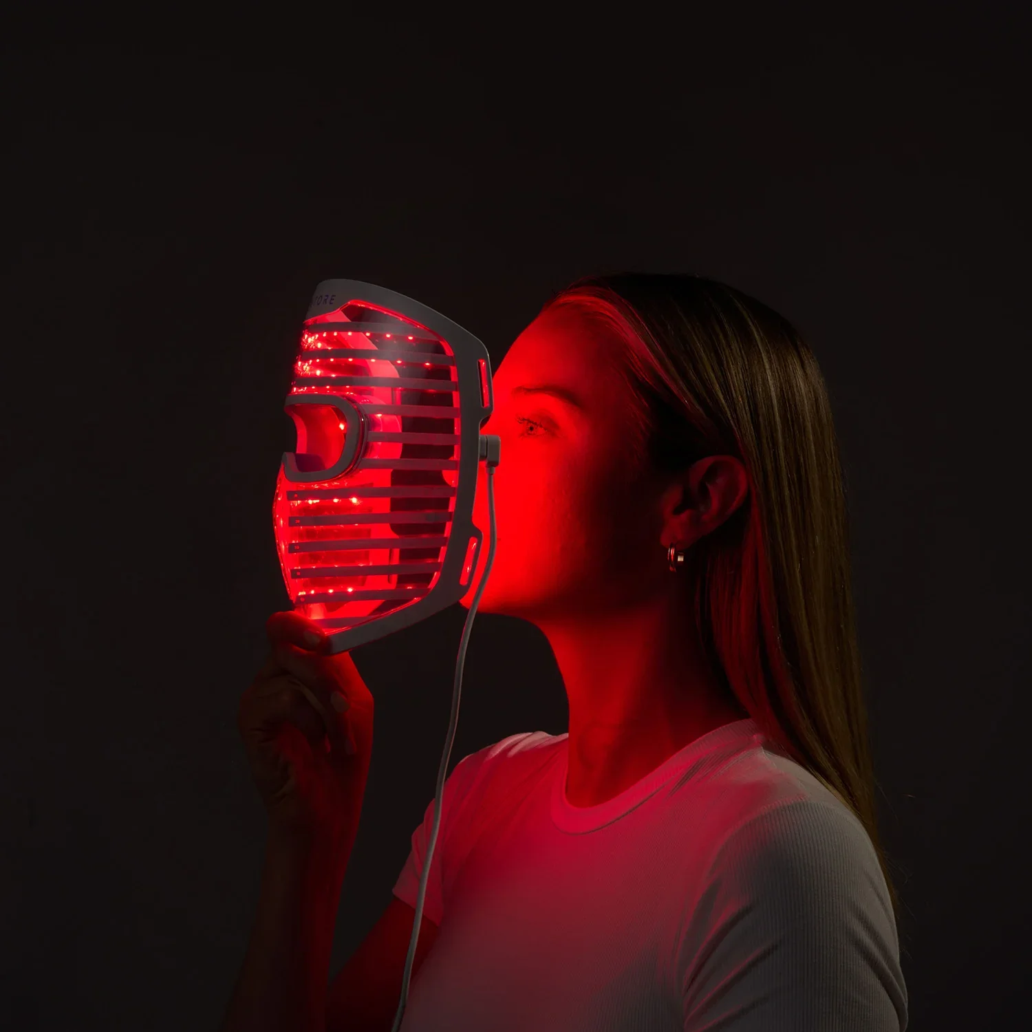 A woman holds a red light mask therapy device close to her face in a dark setting.