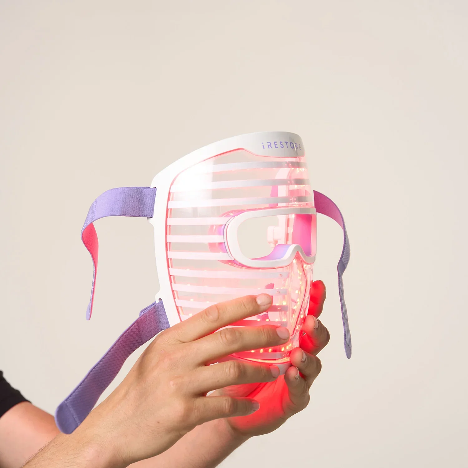 A person holding a pink and white red light mask with purple straps against a plain background.