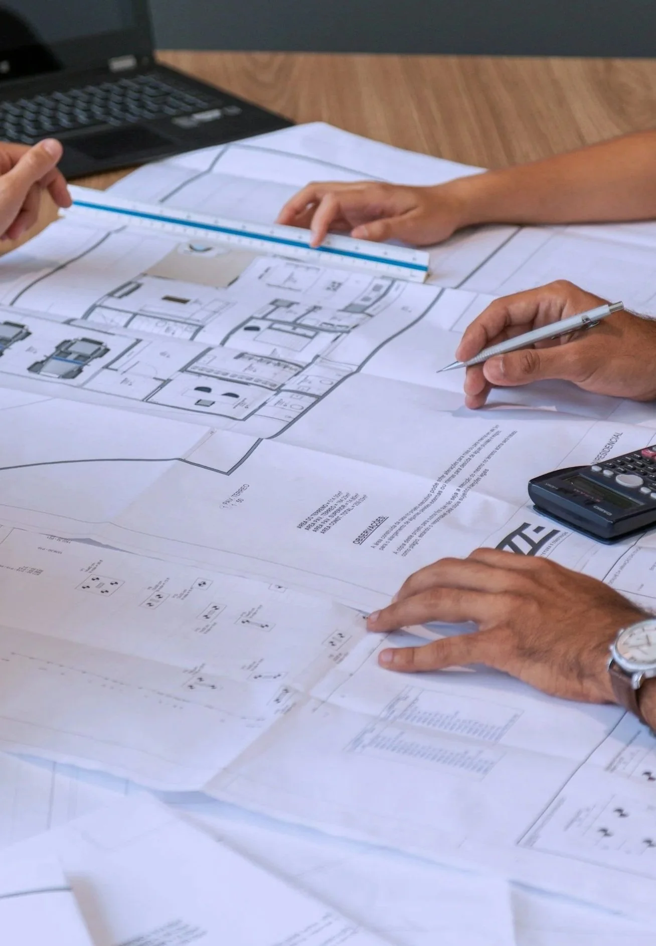 Two people reviewing architectural blueprints at a desk, with a laptop and calculator nearby.