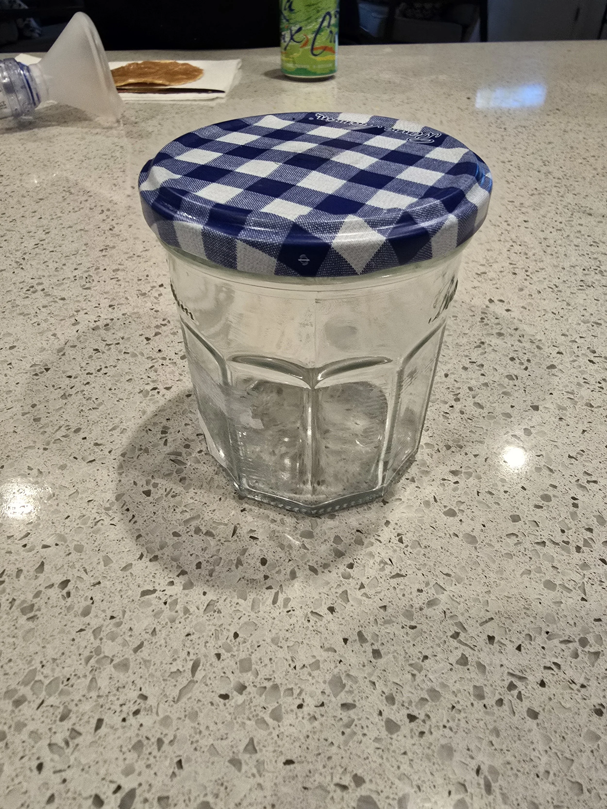 A clear glass jar with a blue and white checkered fabric lid on a speckled beige table. In the background, a can, a bottle, and some paper are visible.