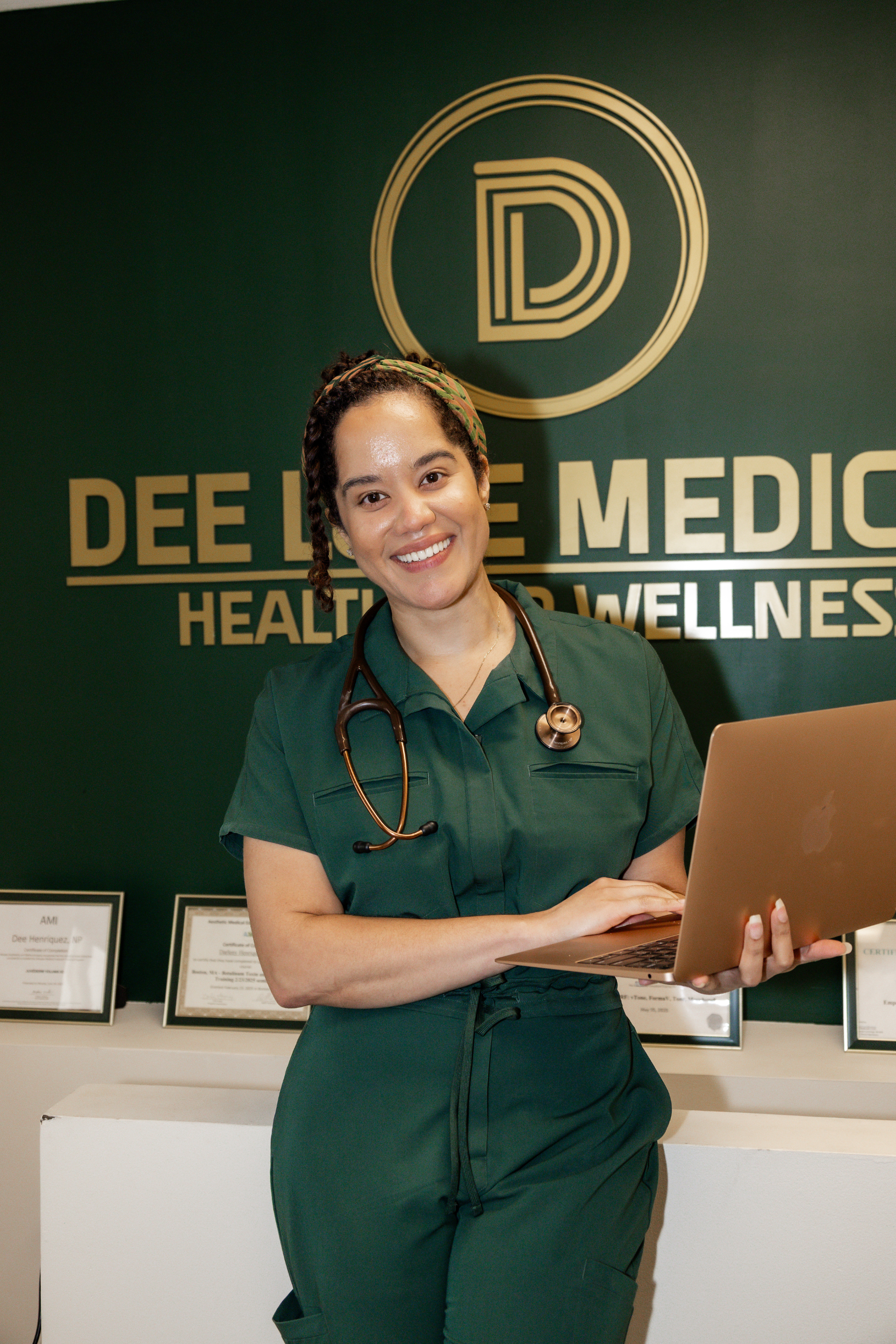 A young female healthcare professional with a stethoscope around her neck, wearing green scrubs, holding a laptop, and smiling, standing in front of a sign that says "DEE