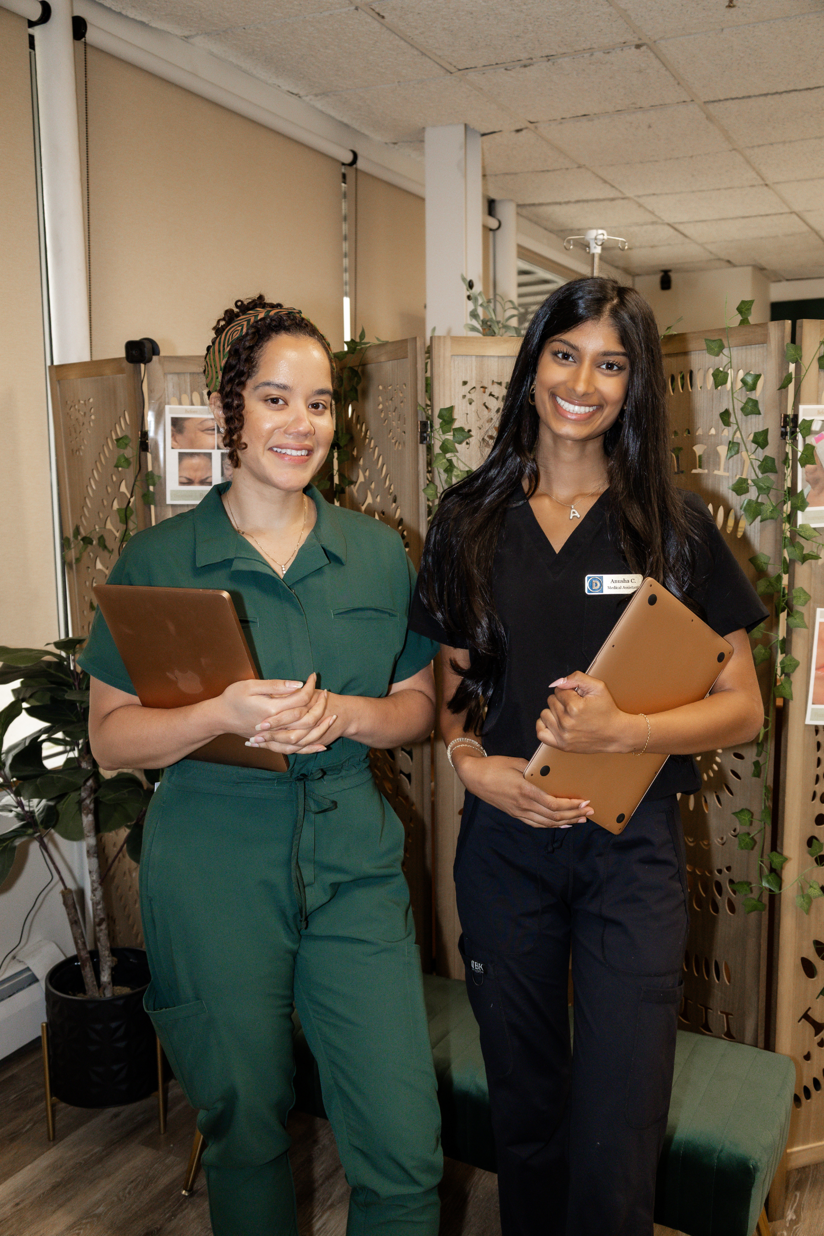 Two women standing together, holding tablets, smiling, in an indoor setting with plants and a wooden divider in the background.