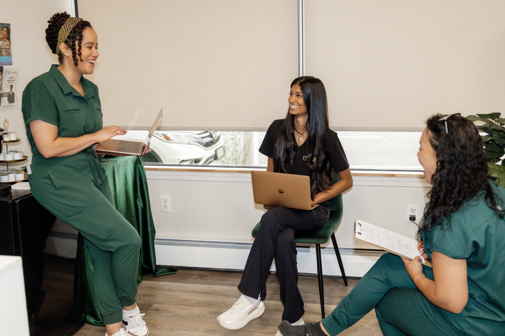 A diverse group of women engaged in a friendly discussion in a well-lit room. One woman in green scrubs is standing and talking, while three others are sitting and listening, with one holding a clipboard and another working on a laptop.