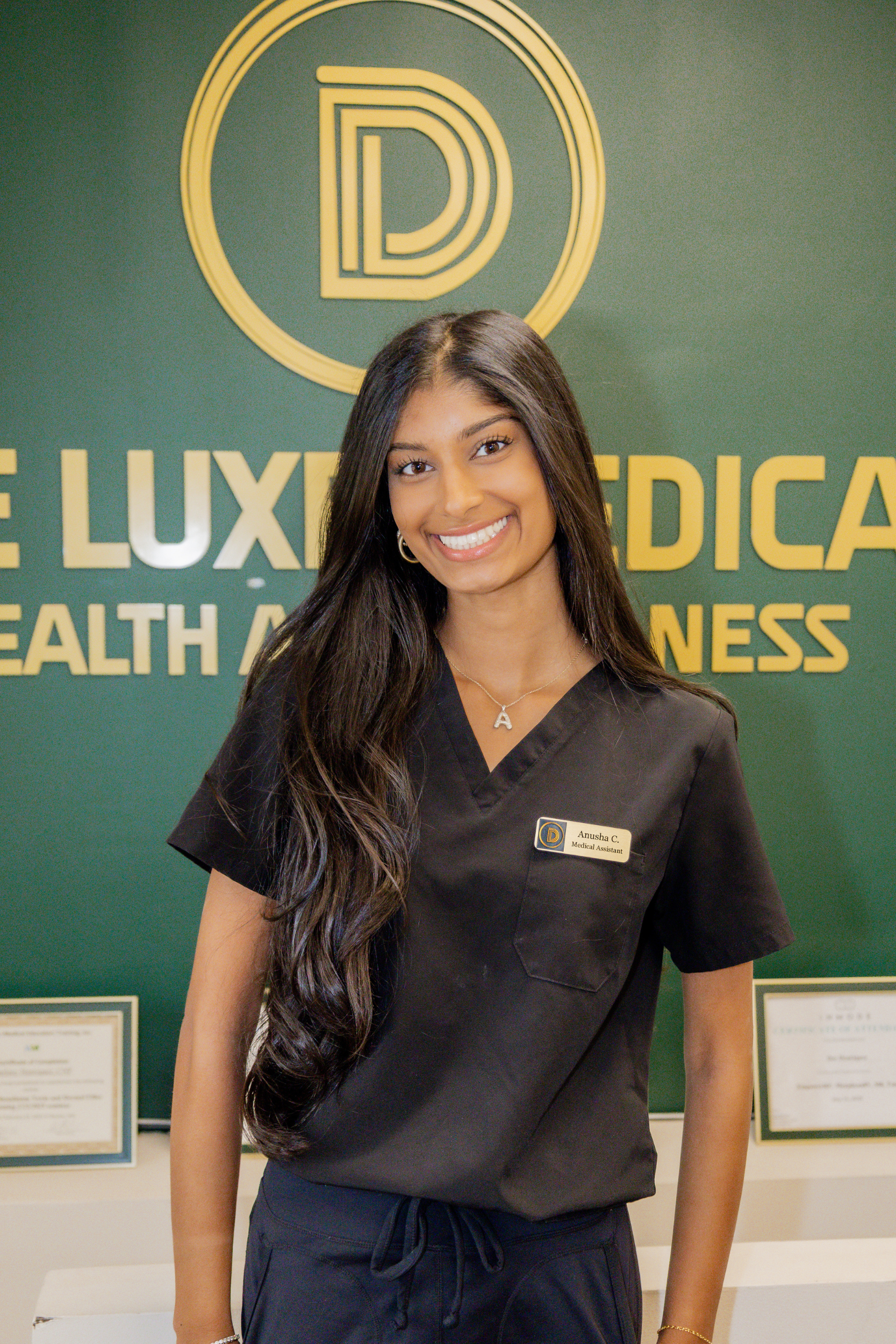 A young woman with long dark hair, smiling, in black medical scrubs with a name tag that reads "Anusha C. Medical Assistant," standing in front of a green background with the words "LUXURY MEDICAL" and other text, along with a circular logo containing a stylized letter "D."