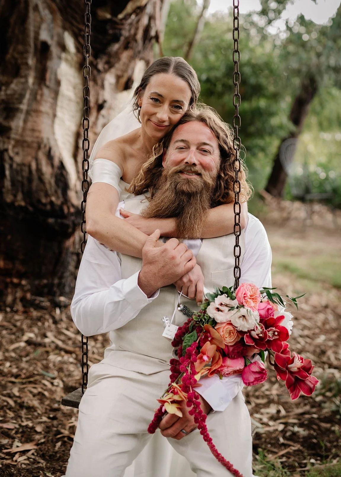 A couple in wedding attire sitting on a swing outdoors, with the woman embracing the man from behind and holding a colorful bouquet of flowers.