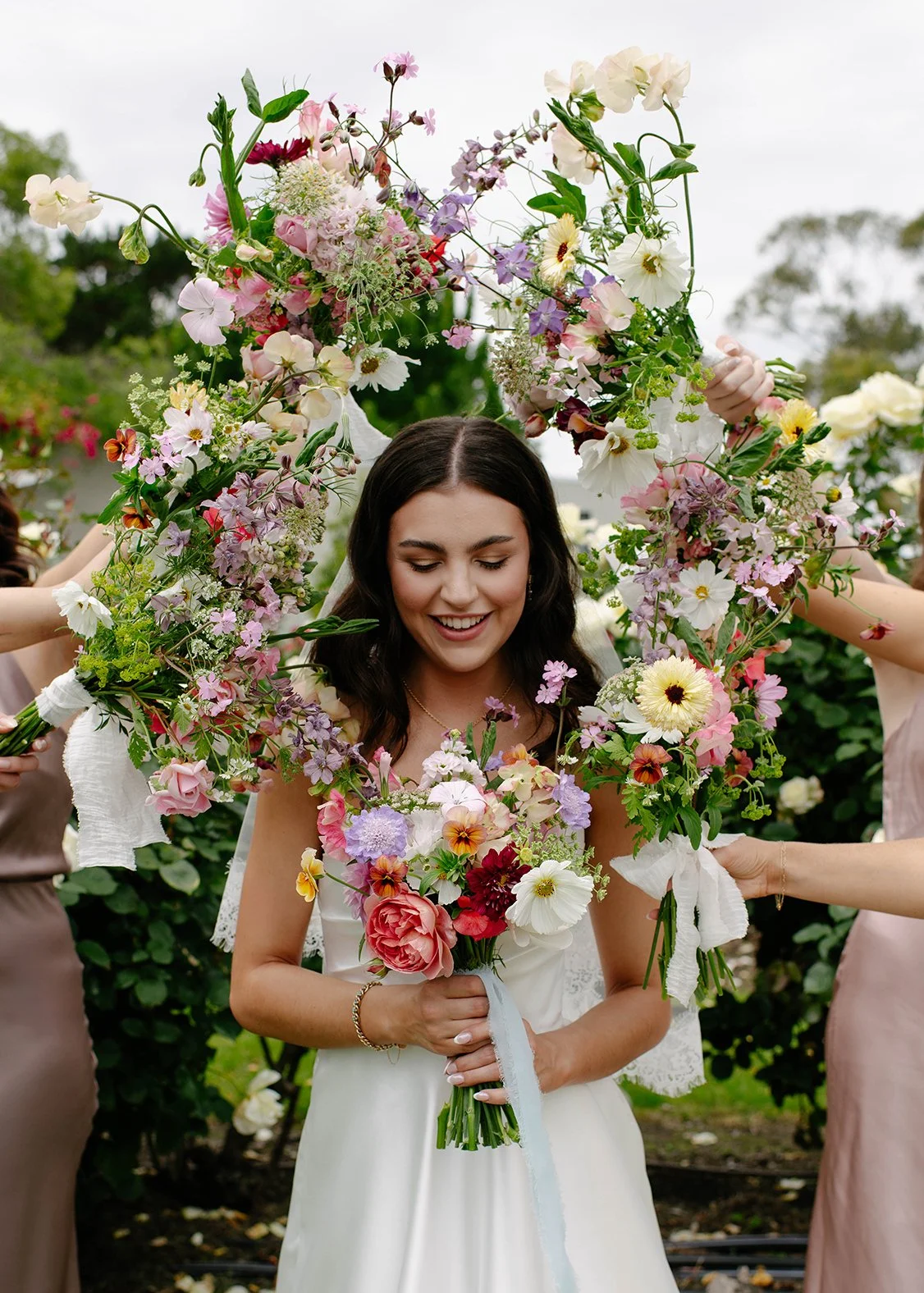 Young woman in white dress holding bouquet of pink, white, and purple flowers, surrounded by others holding floral arrangements, outdoors with greenery and cloudy sky.