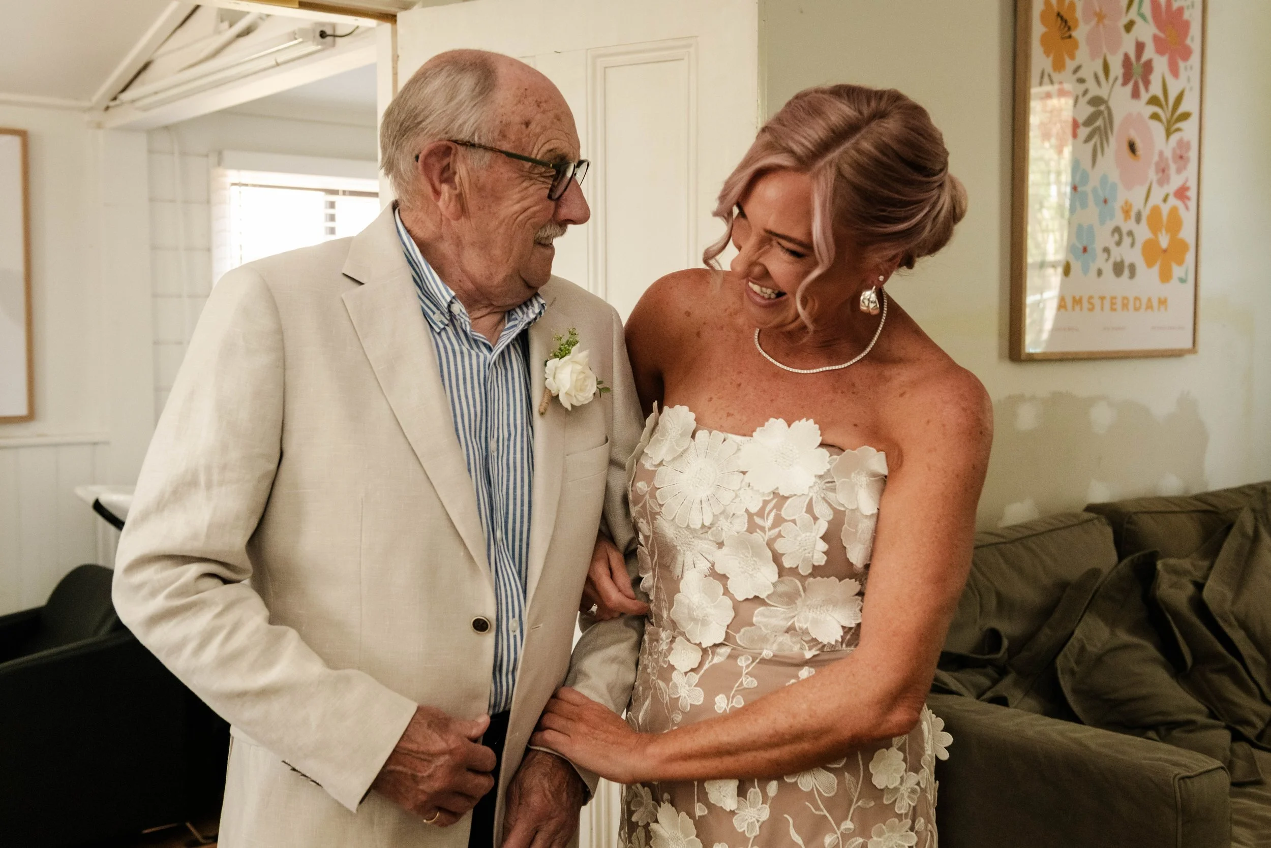 Bride in floral strapless wedding dress smiling with elderly family member during wedding morning preparations.