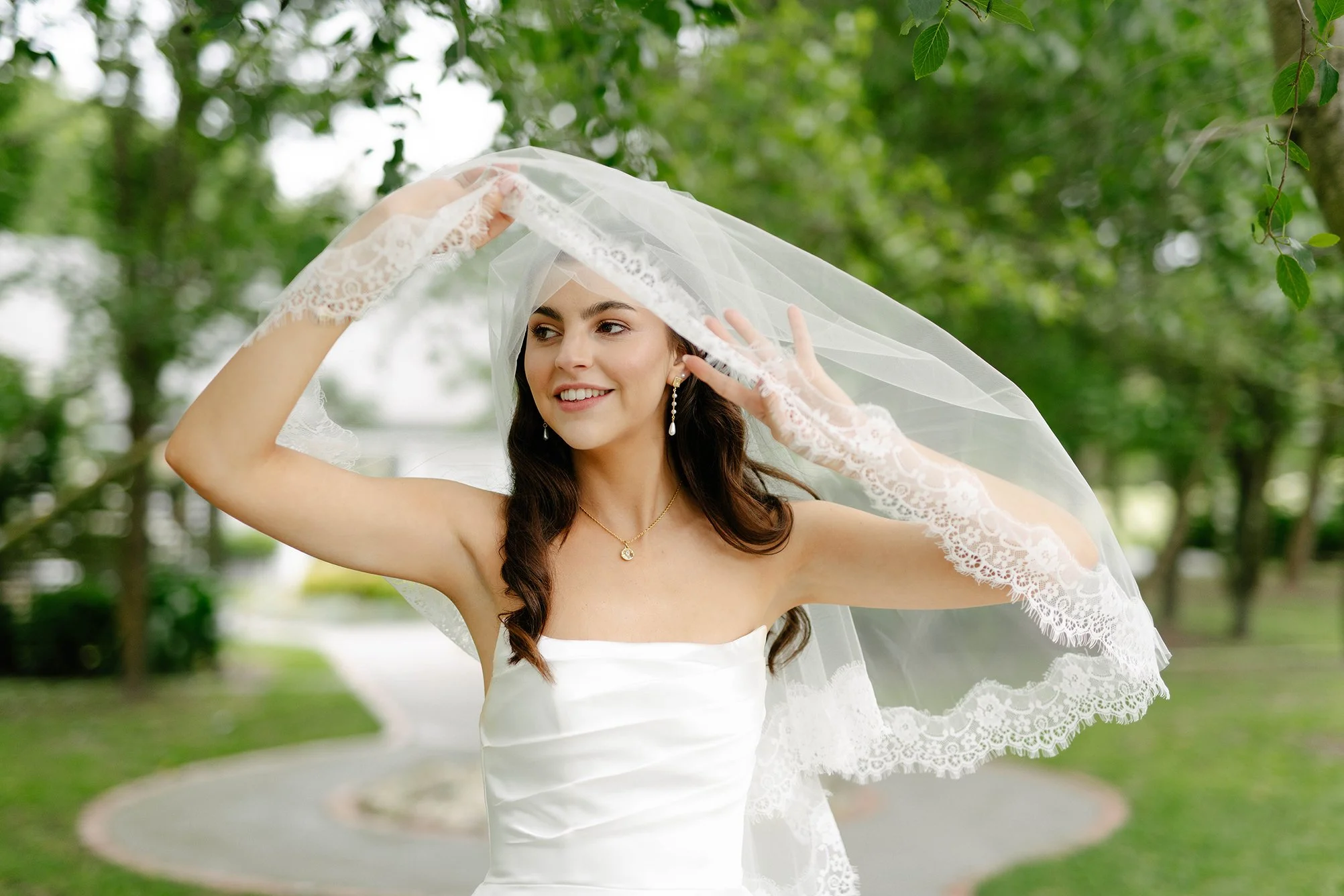 A bride in a white wedding dress holding her veil in a park with green trees.