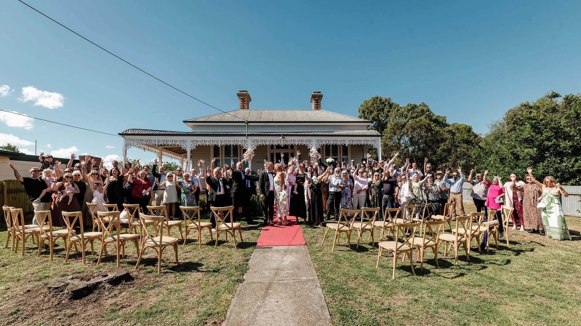 Wedding guests gathered outside a historic-style venue cheering and raising their hands after the ceremony