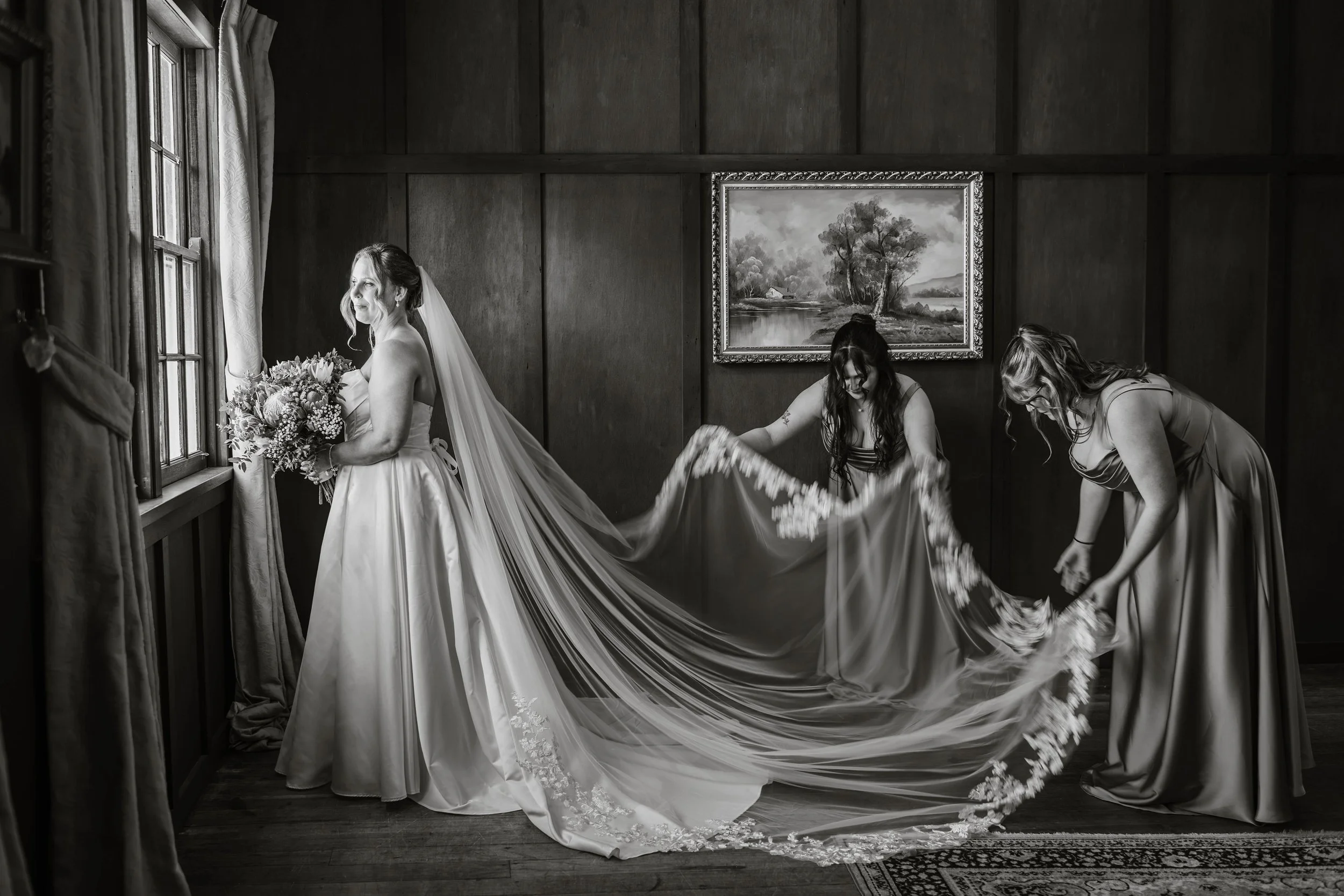 A bride in a wedding dress holding a bouquet looking out a window, with two women adjusting the train of her dress in a wooden-paneled room.
