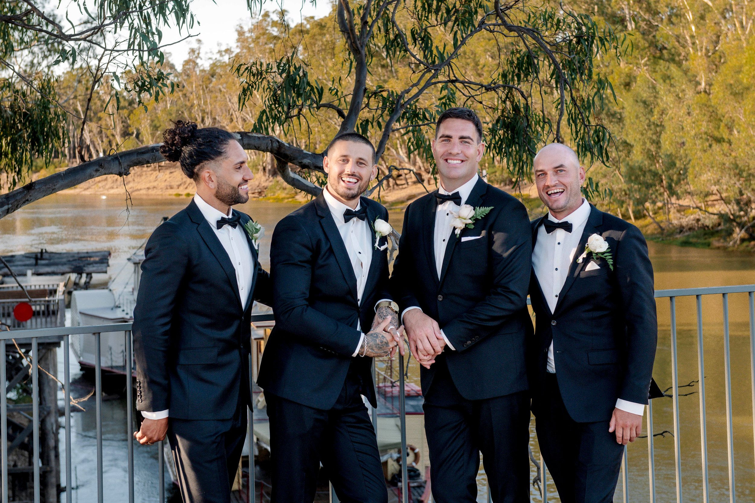 Four men in black tuxedos and bow ties standing outdoors near a river, smiling for a photo, with trees and boats in the background.