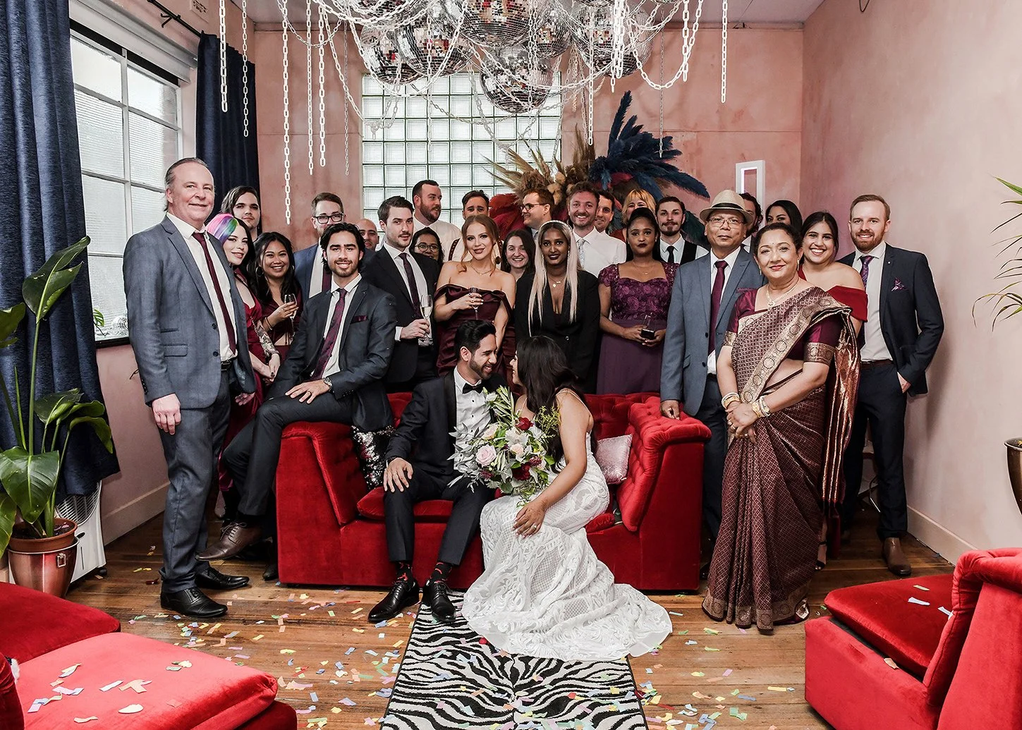 A large group of people at a wedding reception, with a bride and groom sitting on a red velvet sofa at the center, surrounded by guests dressed in formal attire, in a decorated indoor venue with pink walls, large windows, plants, and confetti on the 
