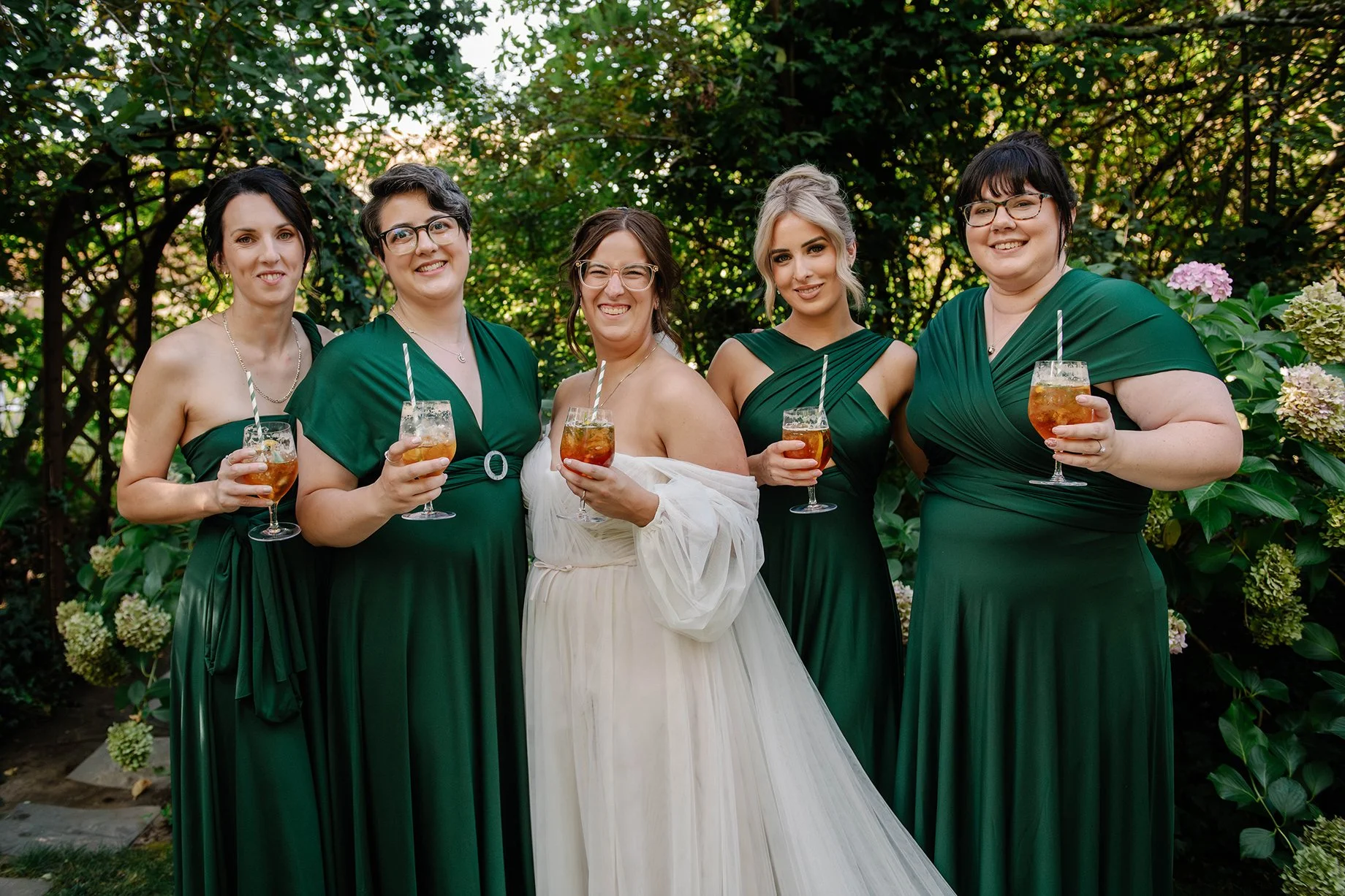 bridal party smiling posed photo with bridesmaid in green dresses holding orange drinks in garden in Bendigo