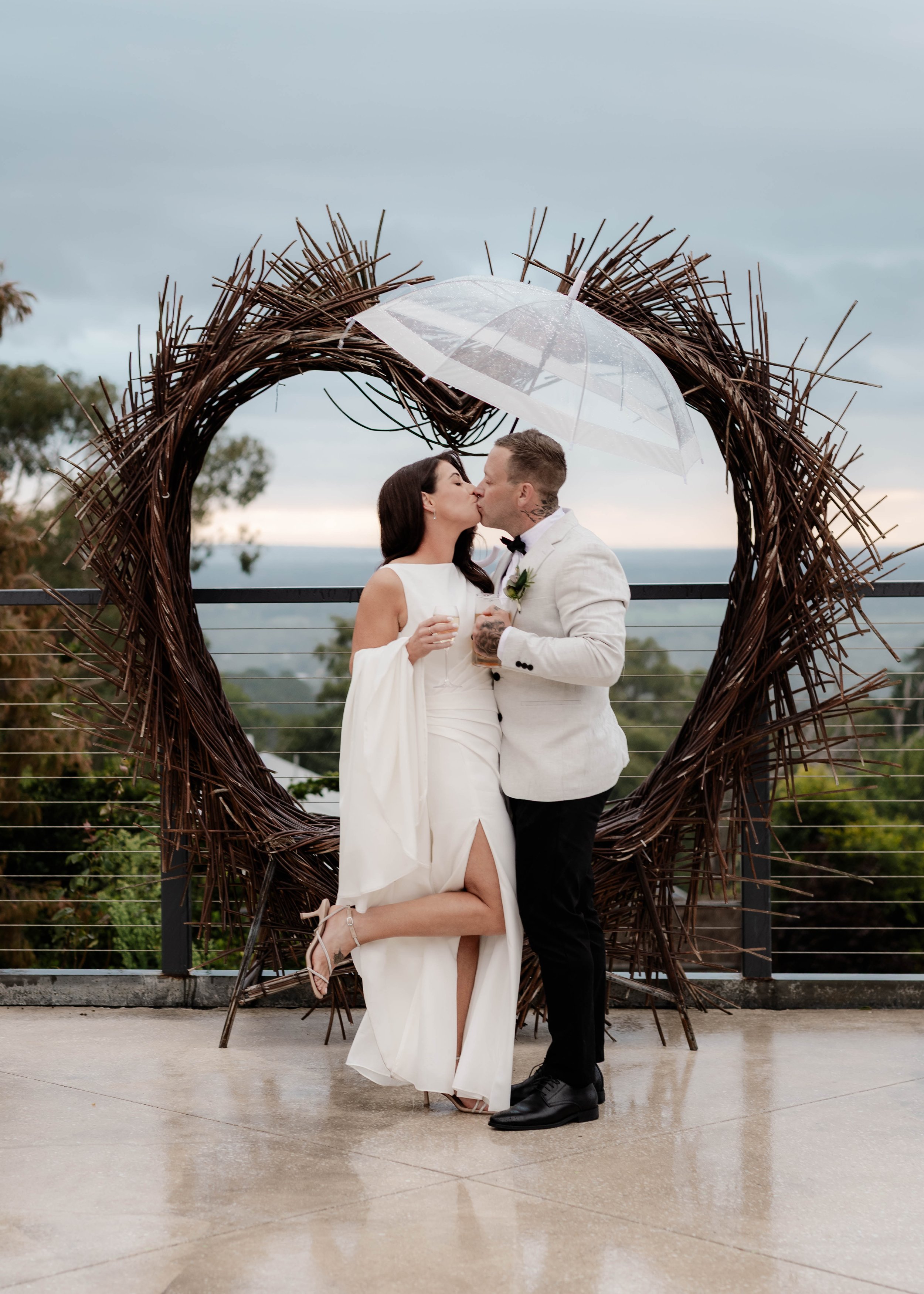 Bride and groom kissing under a clear umbrella in front of a rustic twig arch at Jason's Restaurant Beaconsfield with stormy sky views