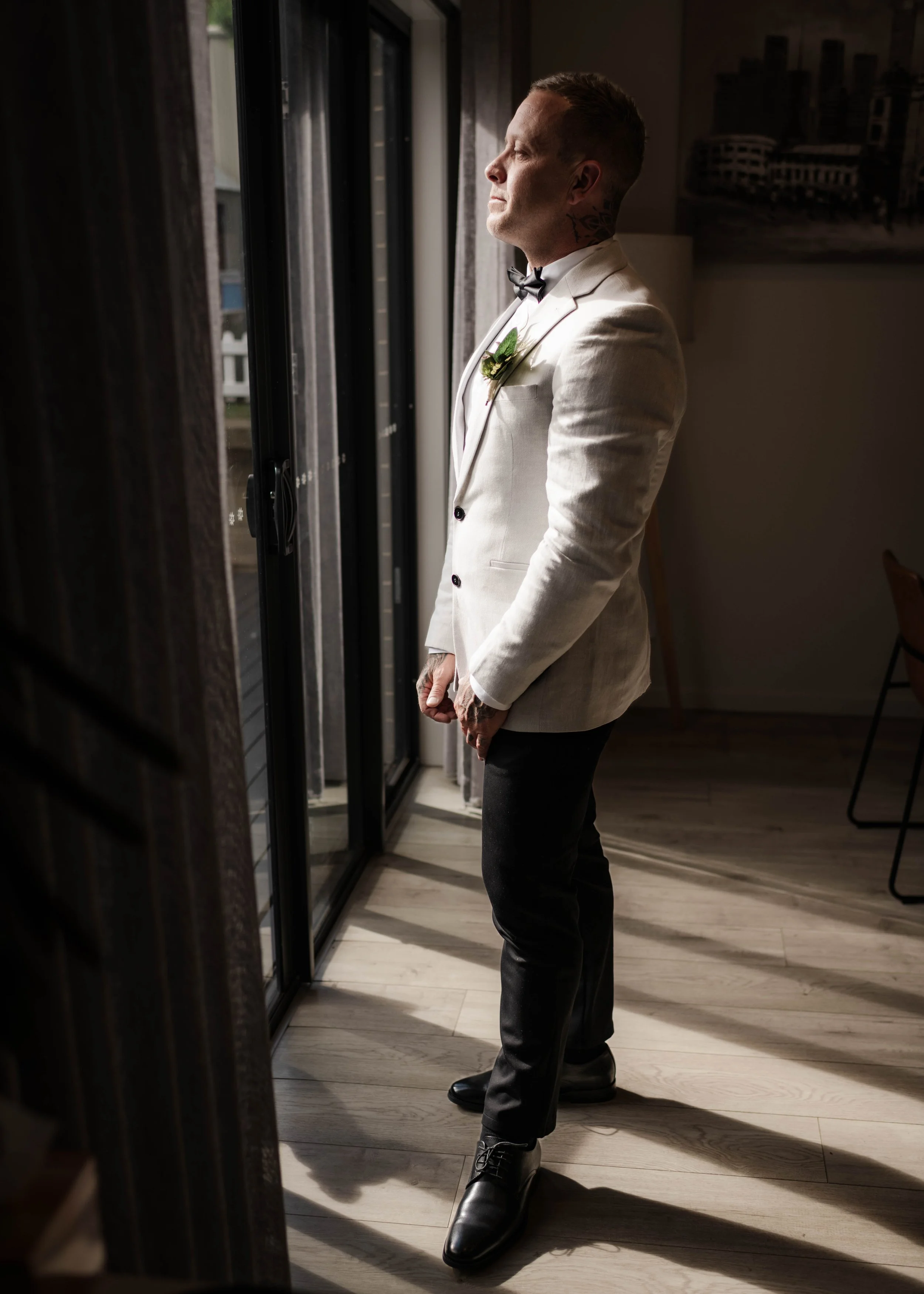 Groom in white jacket and bow tie gazing out a window while getting ready at Jason's Restaurant Beaconsfield