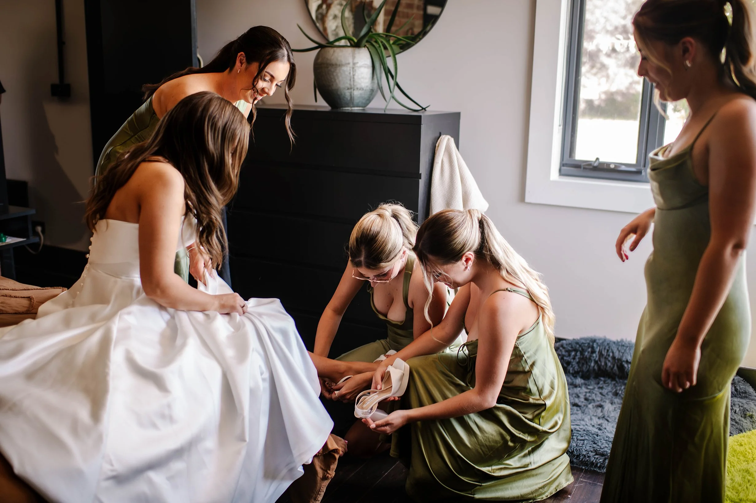 Bridesmaids help bride put on her wedding shoes as she sits on bed in her dress