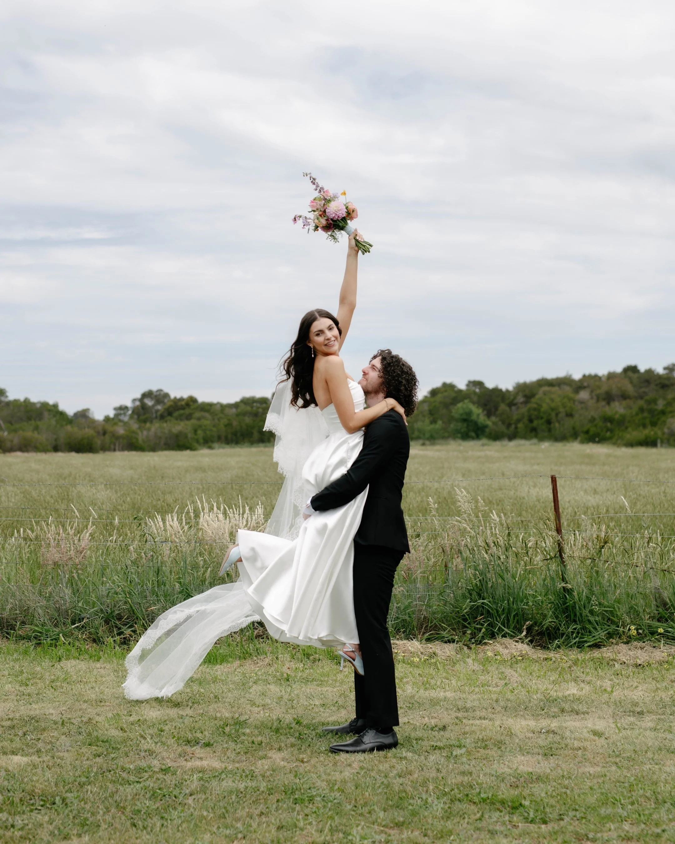 A newlywed couple outdoors in a grassy field, with the groom lifting the bride, who is holding a bouquet, into the air, both smiling, under a cloudy sky before wedding at Daly Waters Mornington.