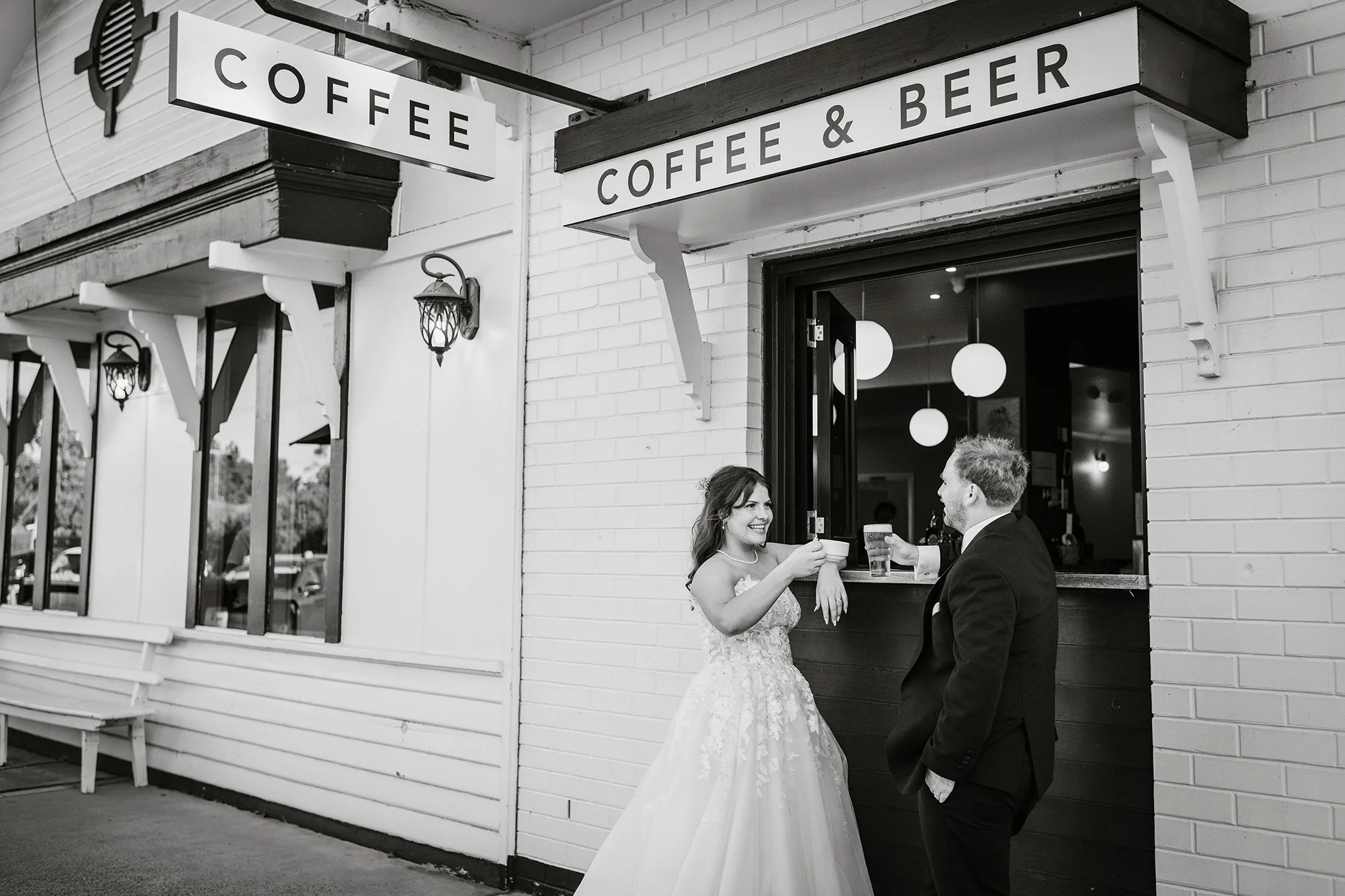 A bride and groom in wedding attire sharing drinks at a coffee and beer stand outside a restaurant, smiling at each other.