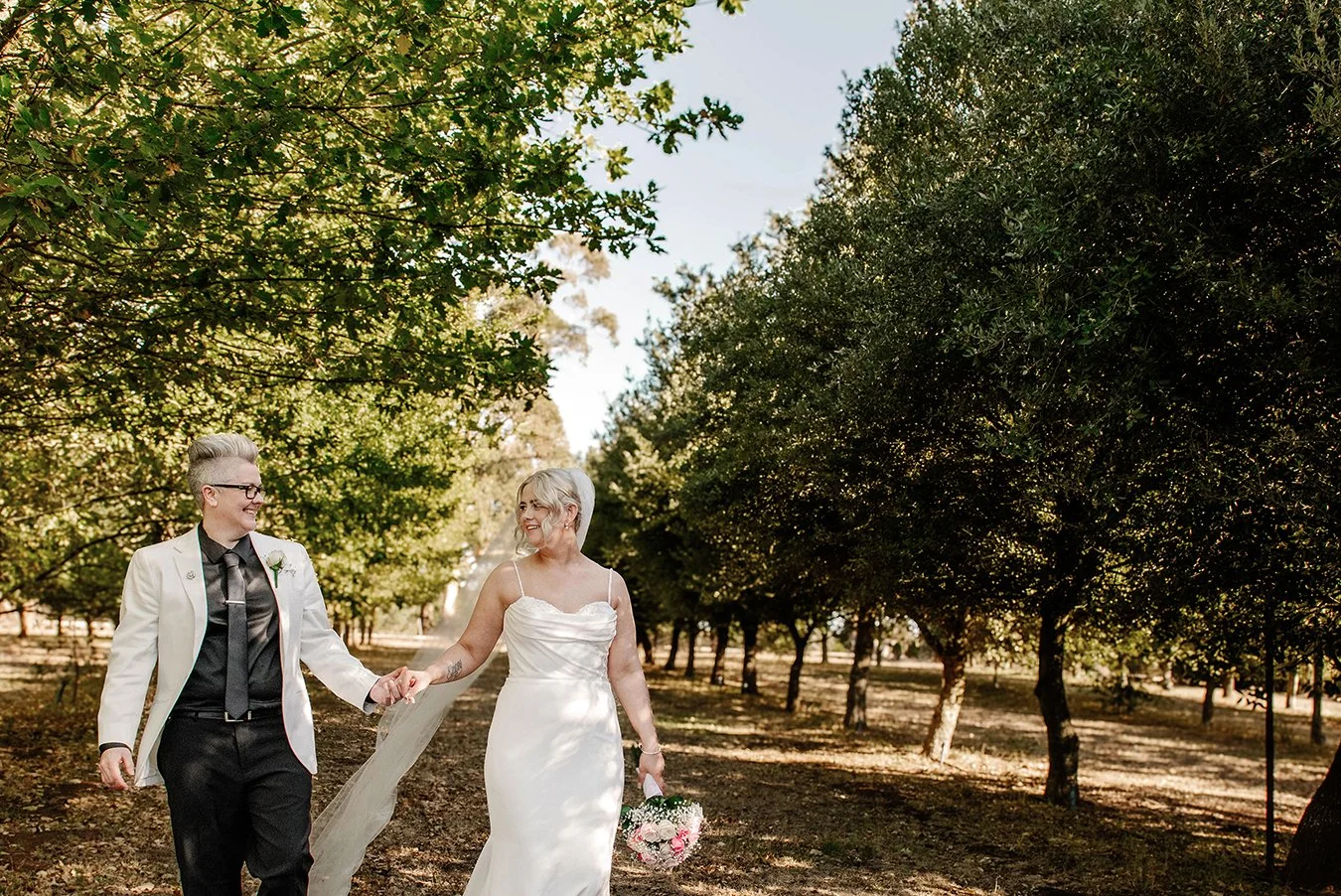 Wedding couple walking hand in hand along a tree-lined orchard path, with one partner holding a bouquet