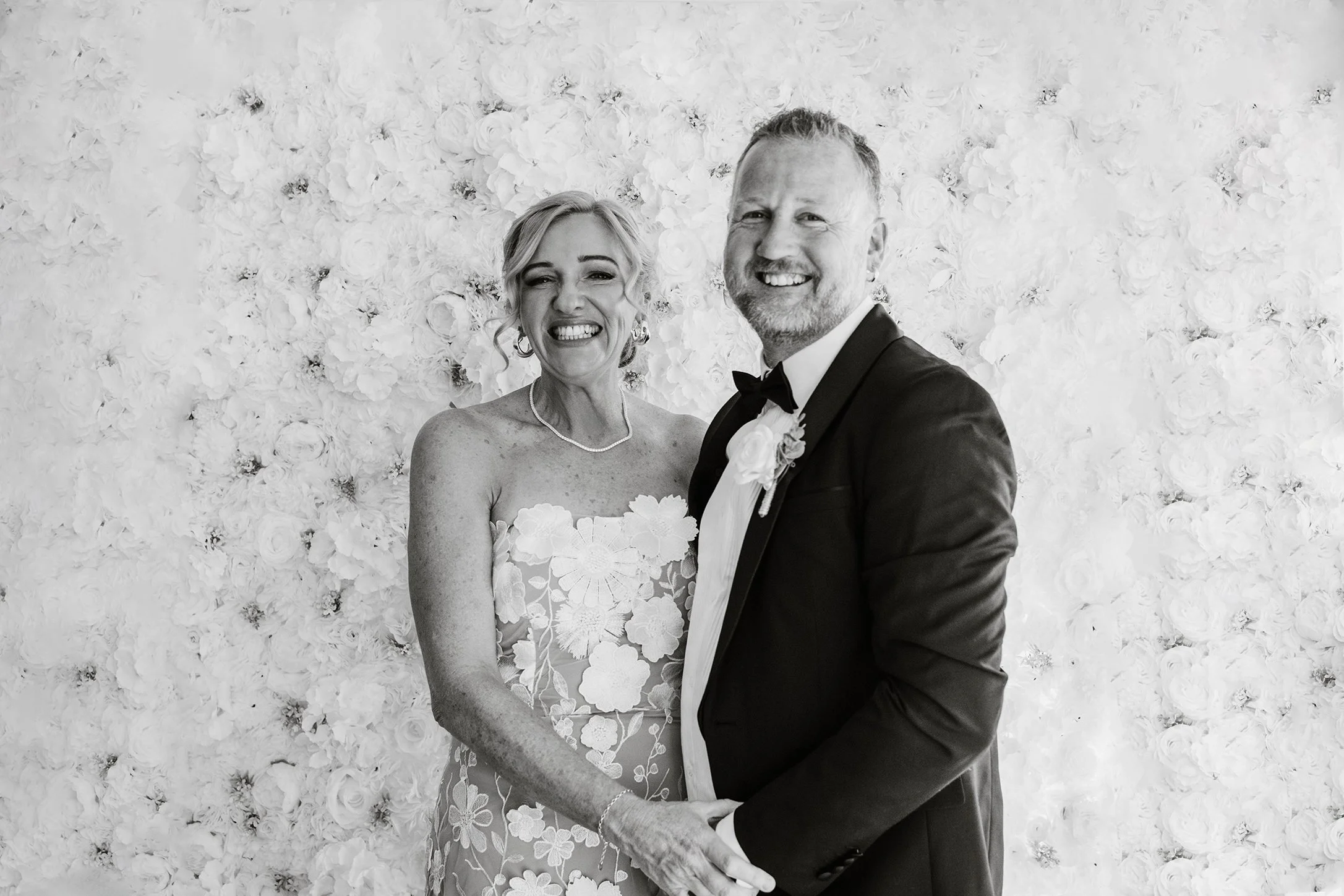 Black and white portrait of the bride and groom holding hands and smiling in front of a floral wall