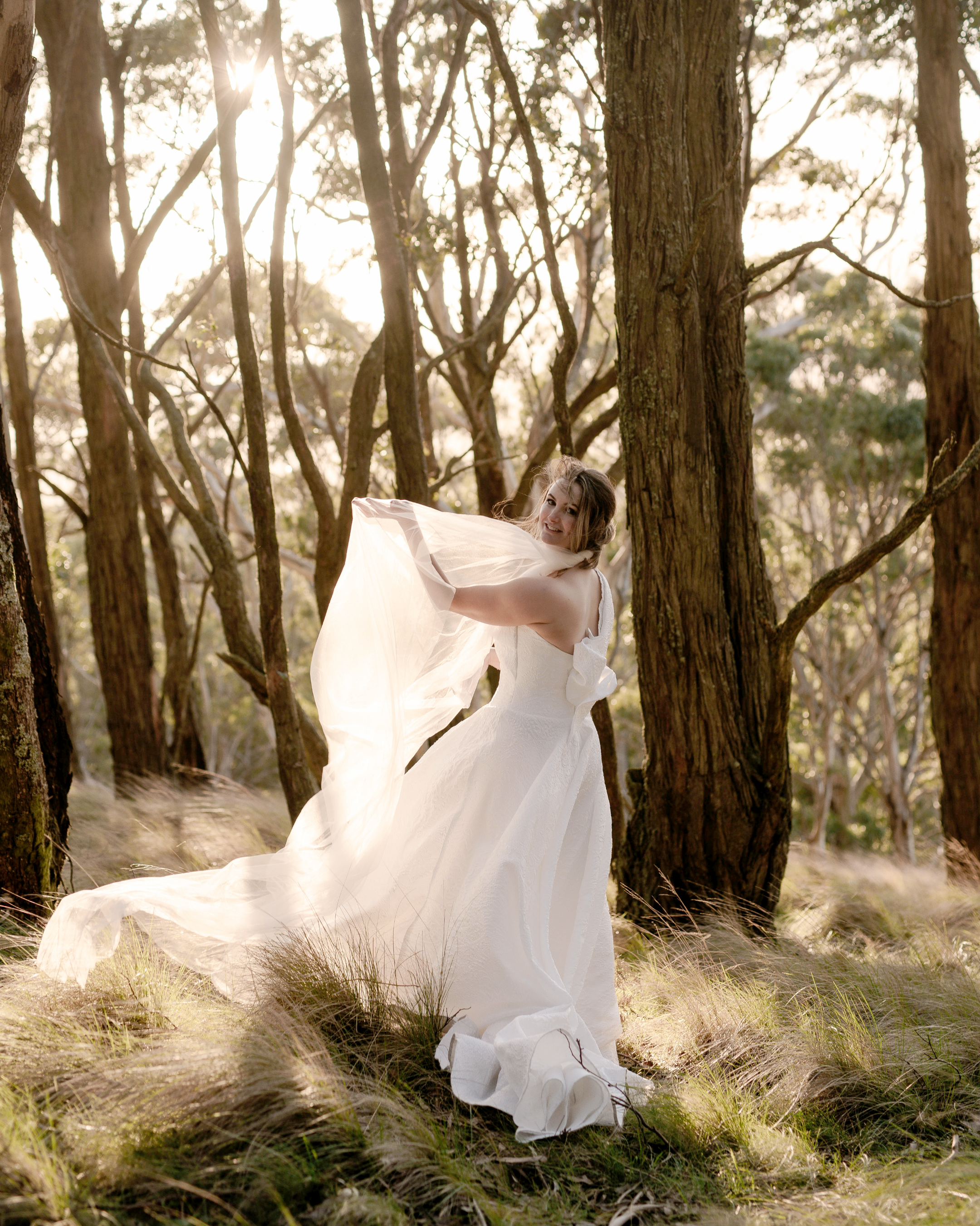 A woman in a white dress and long veil standing in a wooded area with sunlight shining through the trees.