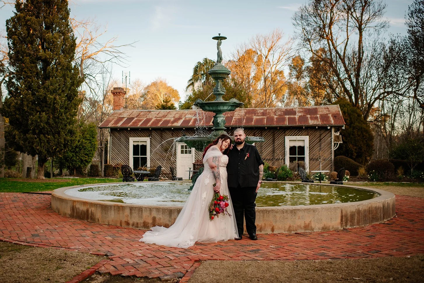 A bride and groom standing together in front of a decorative fountain with a two-tiered sculpture, holding a bouquet of red and pink flowers, outdoors during sunset, with trees and a rustic wooden building in the background.