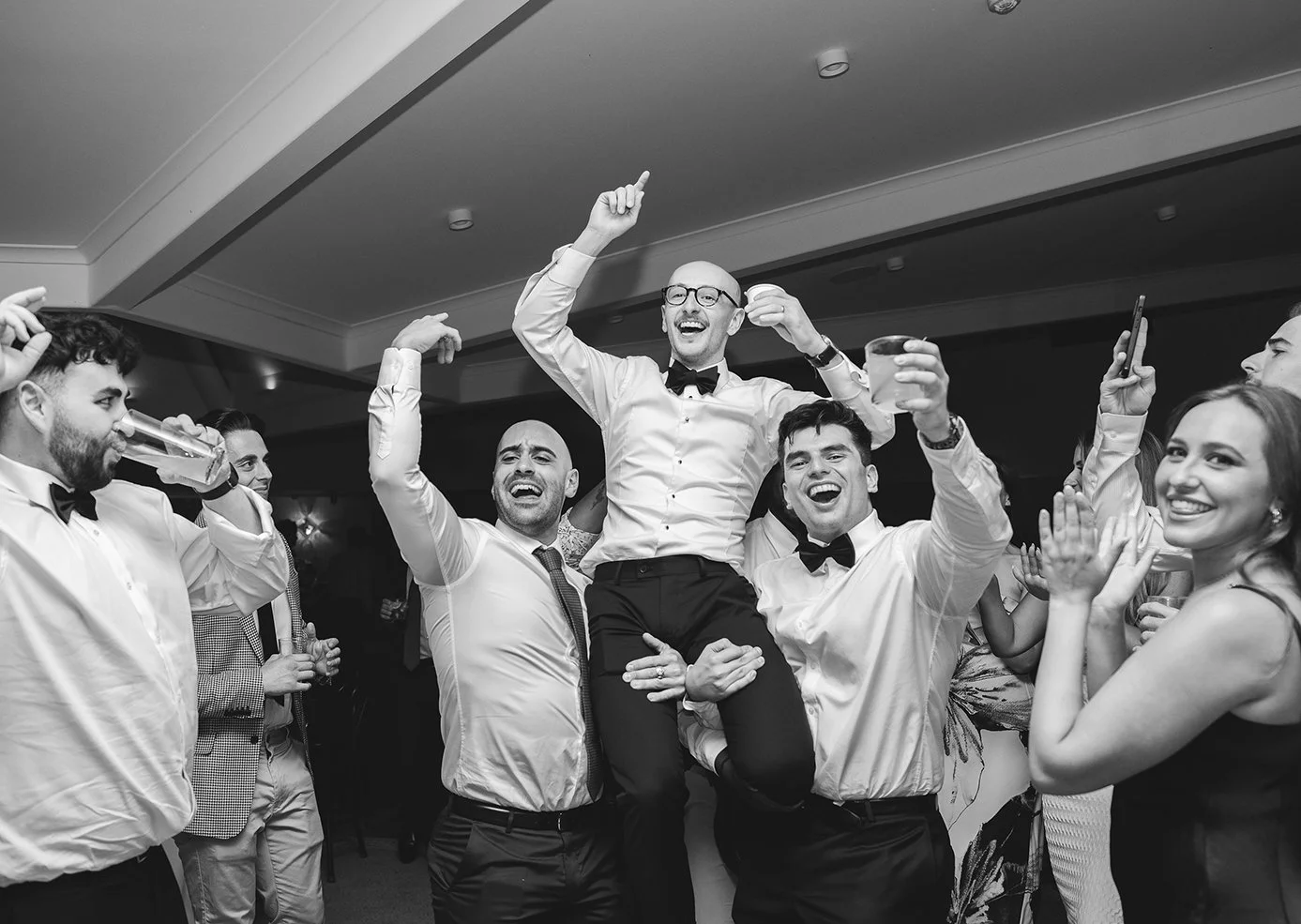 People celebrating at a party with one man being lifted up, smiling, wearing glasses and a bowtie, surrounded by friends with drinks and clapping.