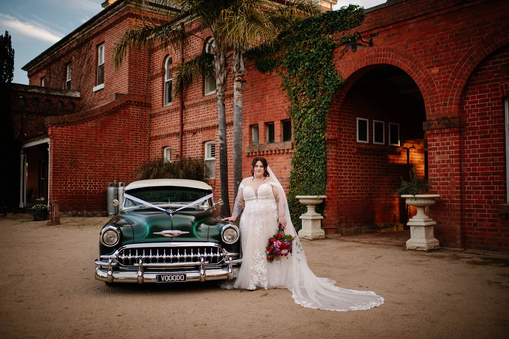 A bride in a white wedding dress standing next to a vintage black and white car with a wedding ribbon in front of a red brick building with palm trees.