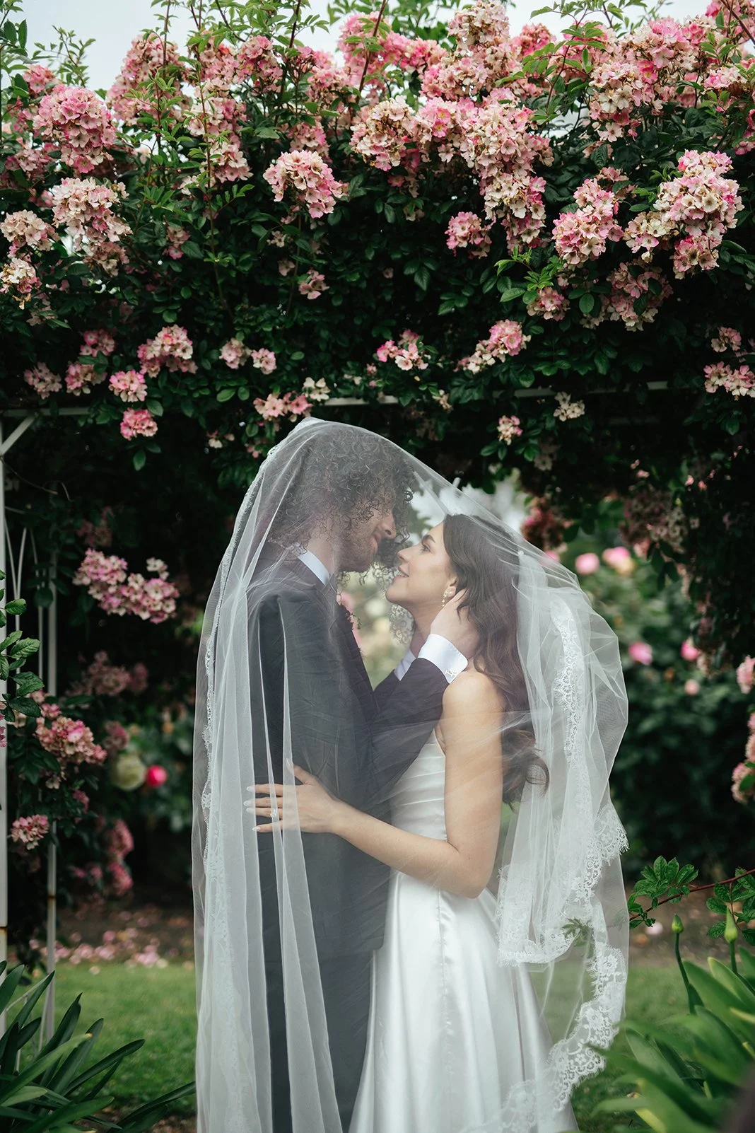 A bride and groom share a romantic moment under a floral arch, with the bride’s veil covering both of them, surrounded by pink flowering bushes.