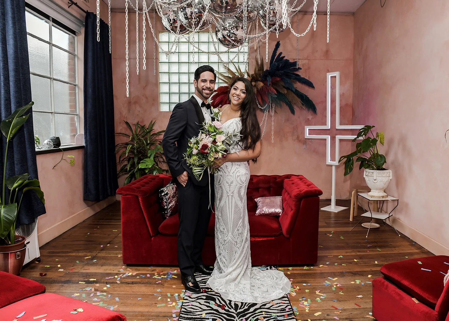 Bride and groom standing on a zebra print rug in a decorated room, smiling, with confetti on the floor, plants, a red velvet sofa, and decorative wall elements in the background.