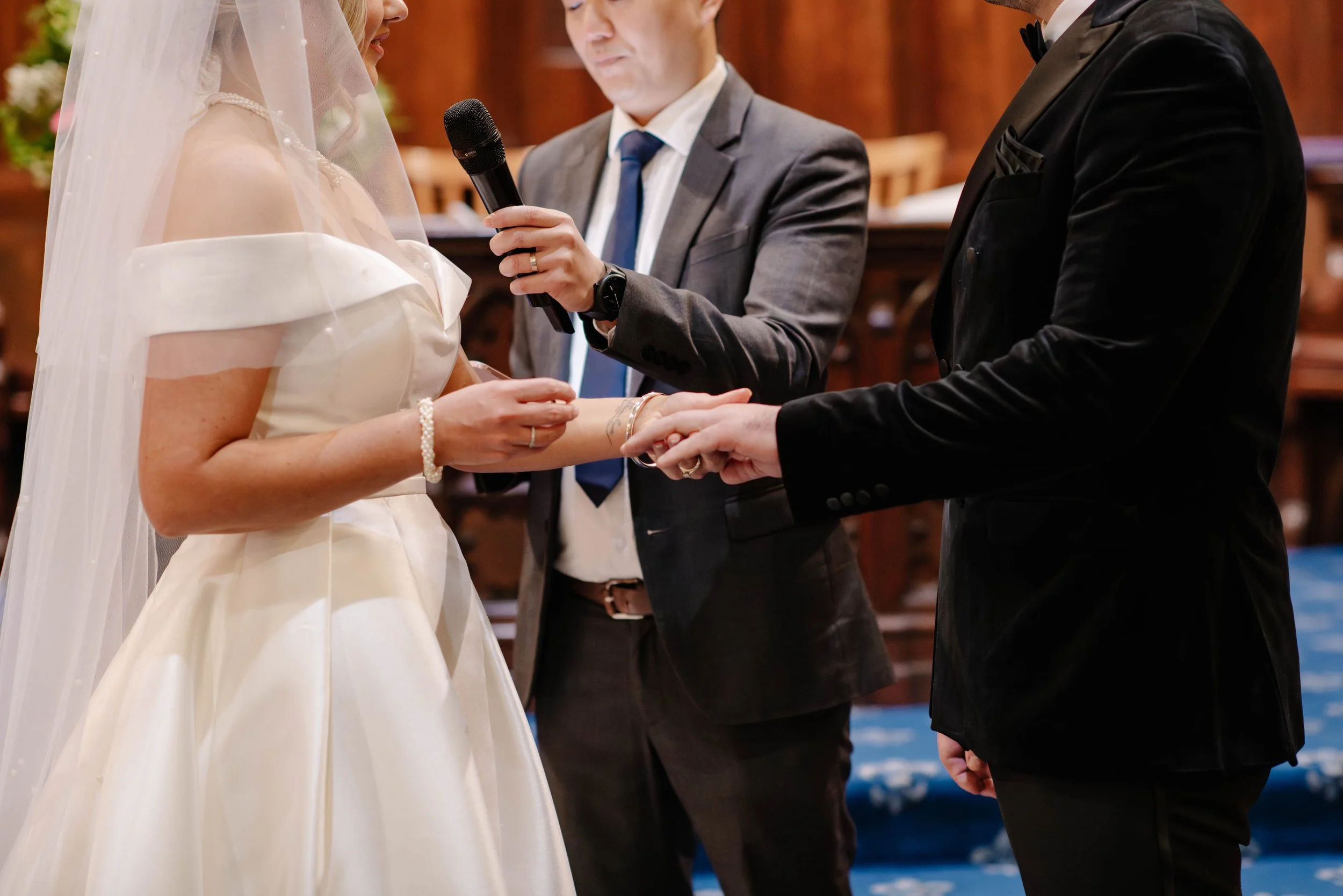 close up view of bride putting on grooms ring as pastor holds a microphone for her