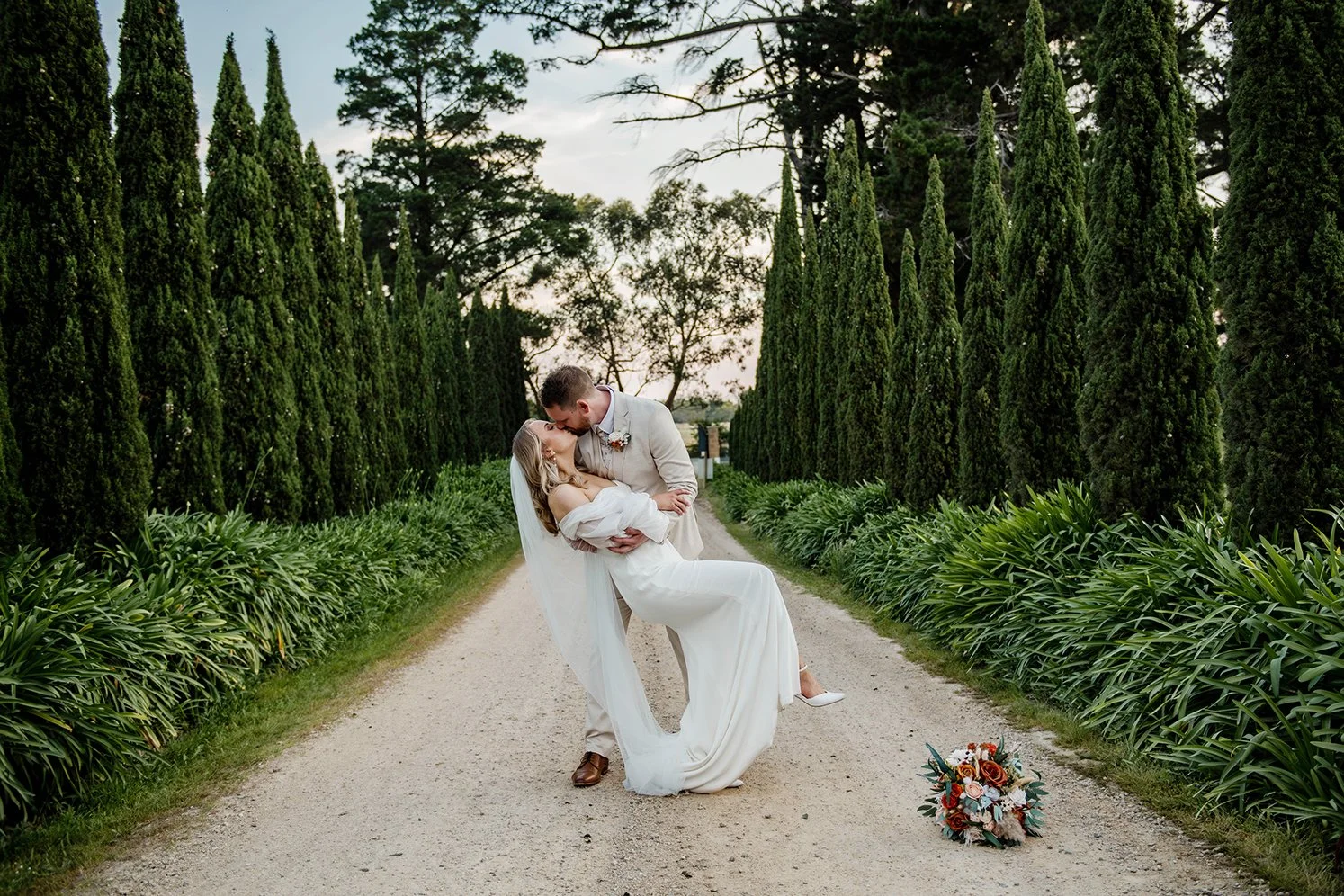 A couple in wedding attire sharing a kiss in a lush garden pathway lined with tall, green trees and low bushes. The woman is being dipped by the man, who is holding her and leaning in to kiss her. A bouquet of flowers is on the ground nearby.