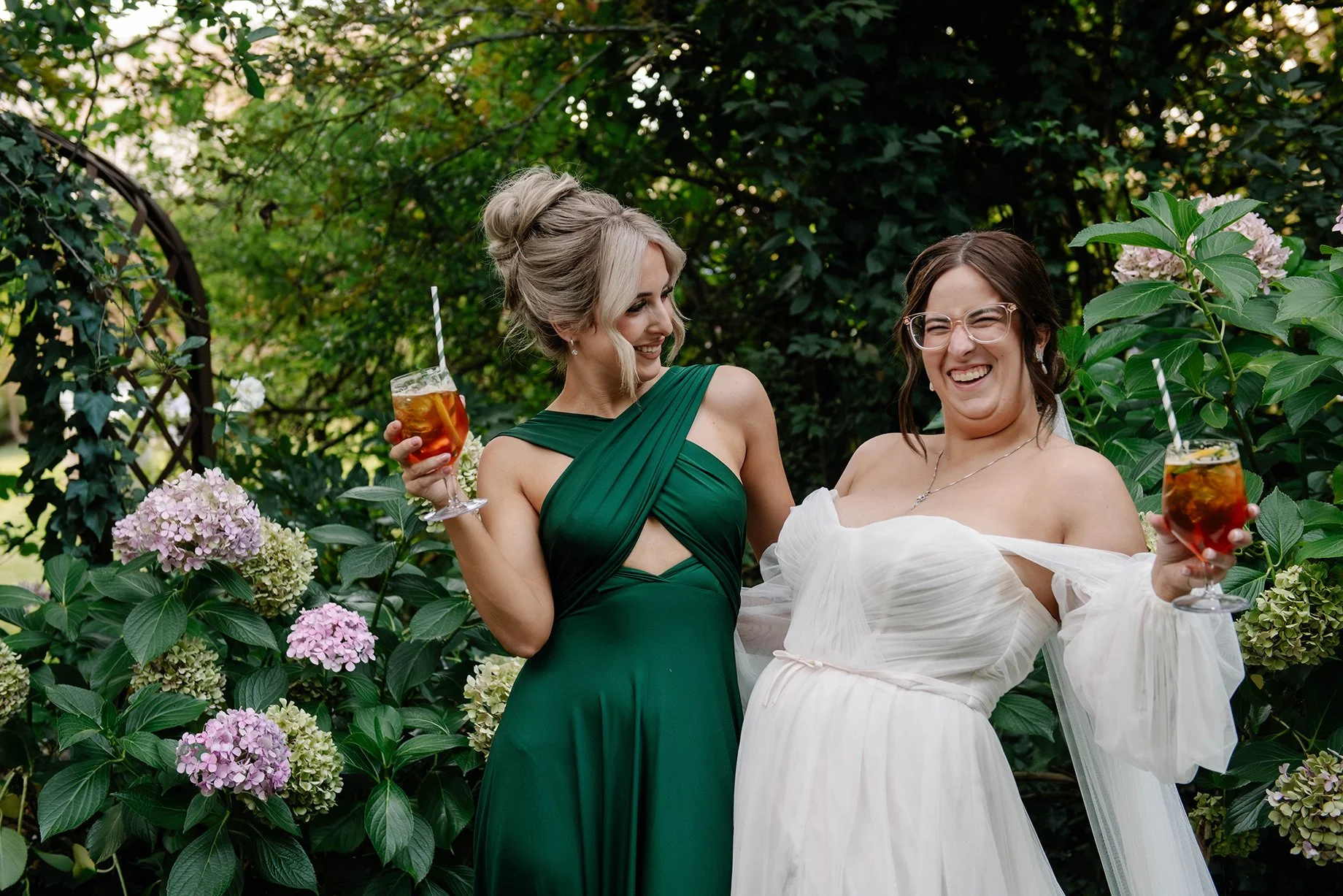 bride and maid of honor posing and smiling with orange drinks in lush green garden in Bendigo