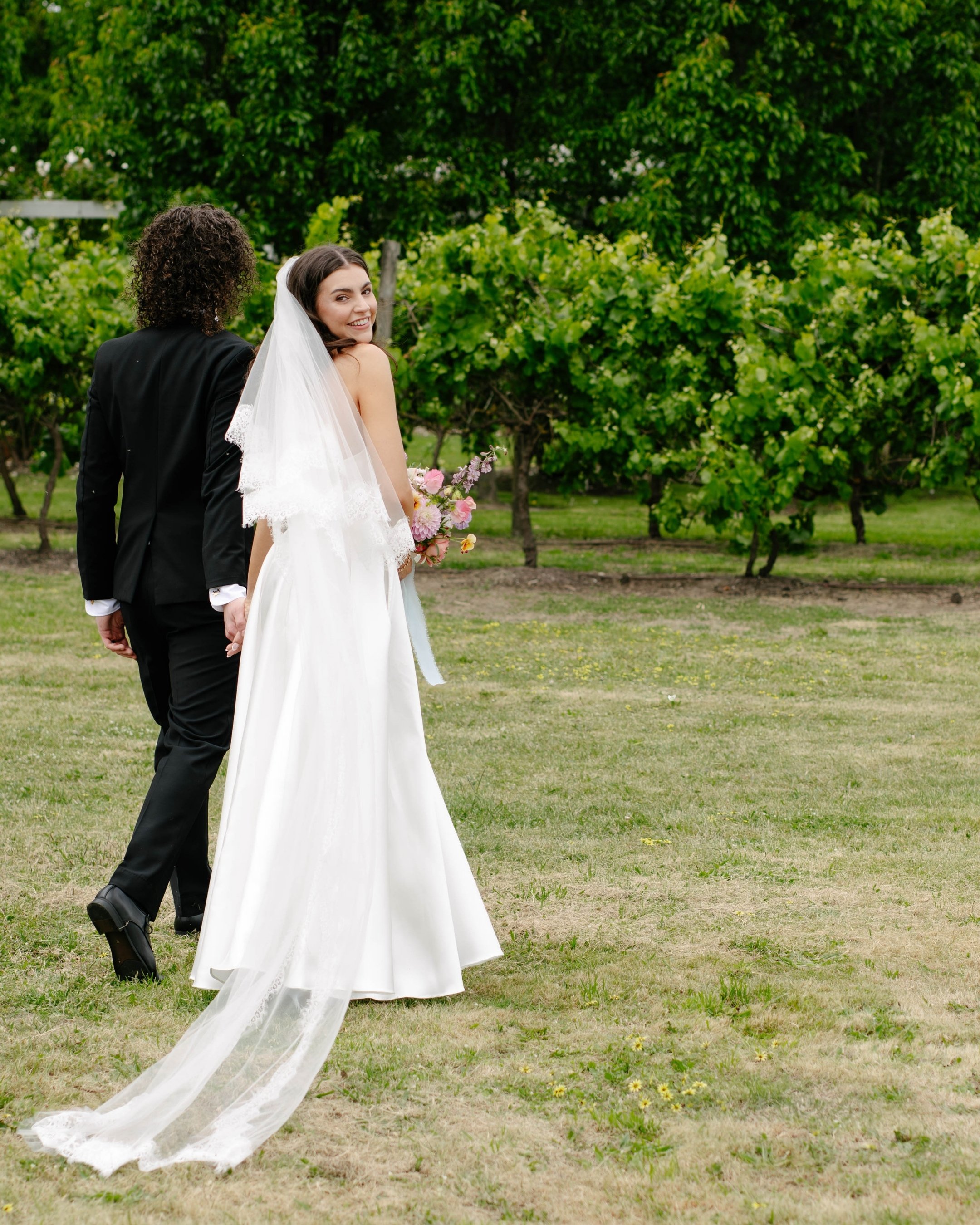 A bride in a white wedding gown and veil holding a bouquet of pink flowers, smiling and looking back over her shoulder, holding hands with a groom dressed in a black suit, walking outdoors on a grassy area with green trees in the background.