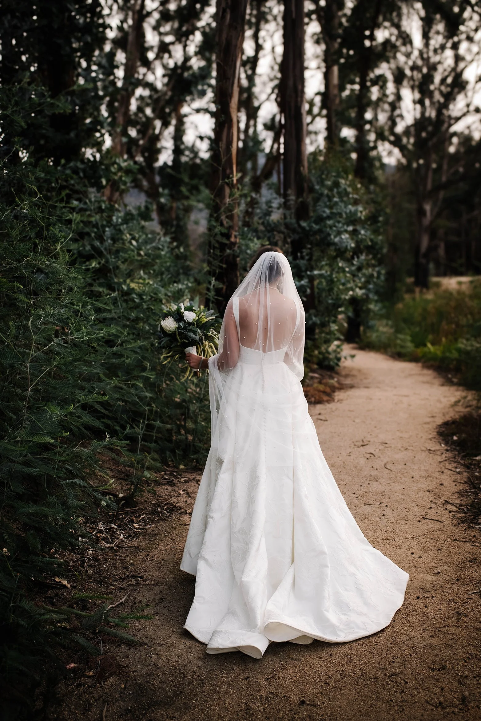 A bride in a white wedding dress and veil holding a bouquet, standing on a dirt path in a forest with tall trees and green foliage.