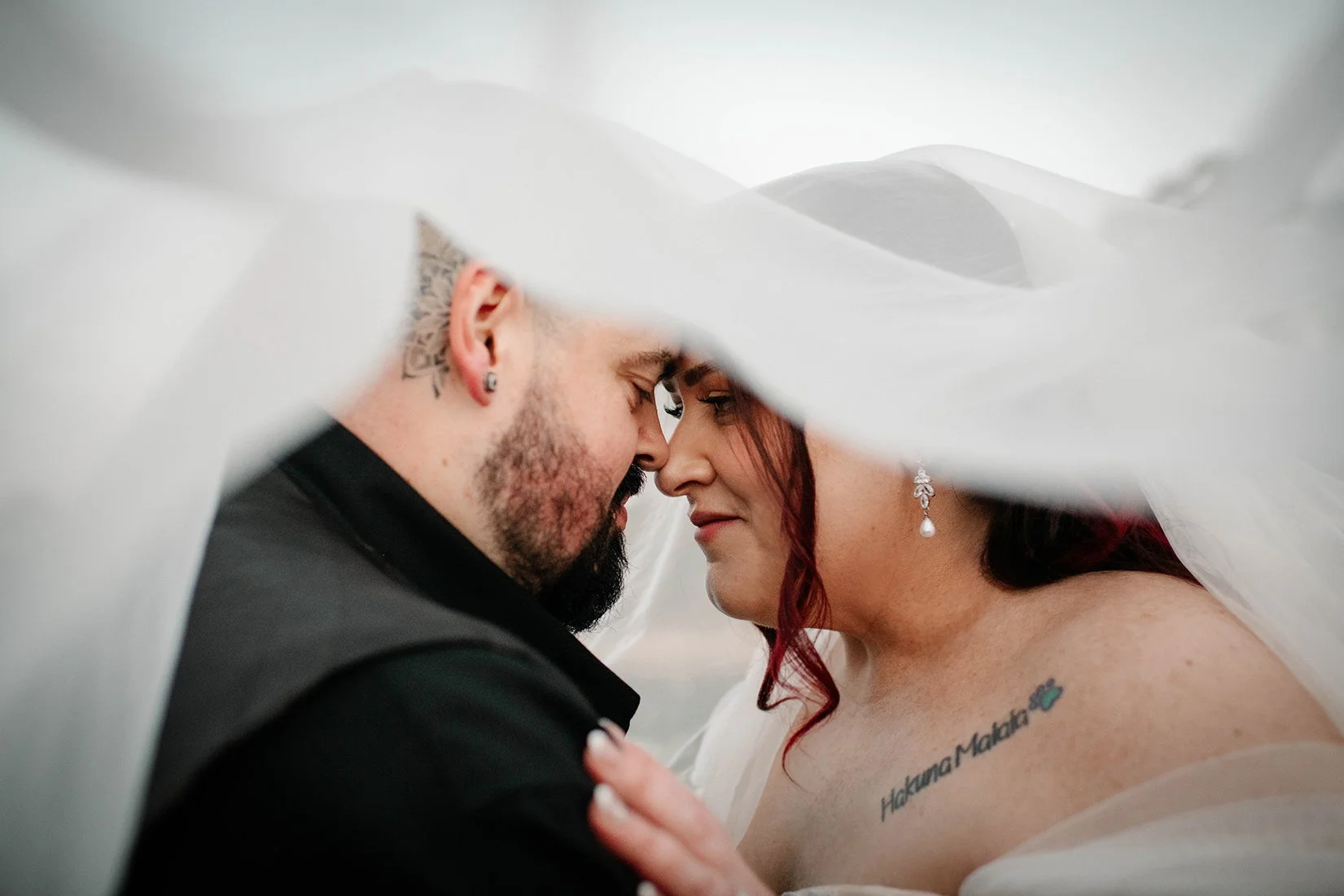 A newlywed couple is captured close under a white veil, touching foreheads, with the groom's face partially visible and the bride's face shown, featuring red hair, earrings, and a tattoo reading 'Hakuna Matata' on her shoulder.