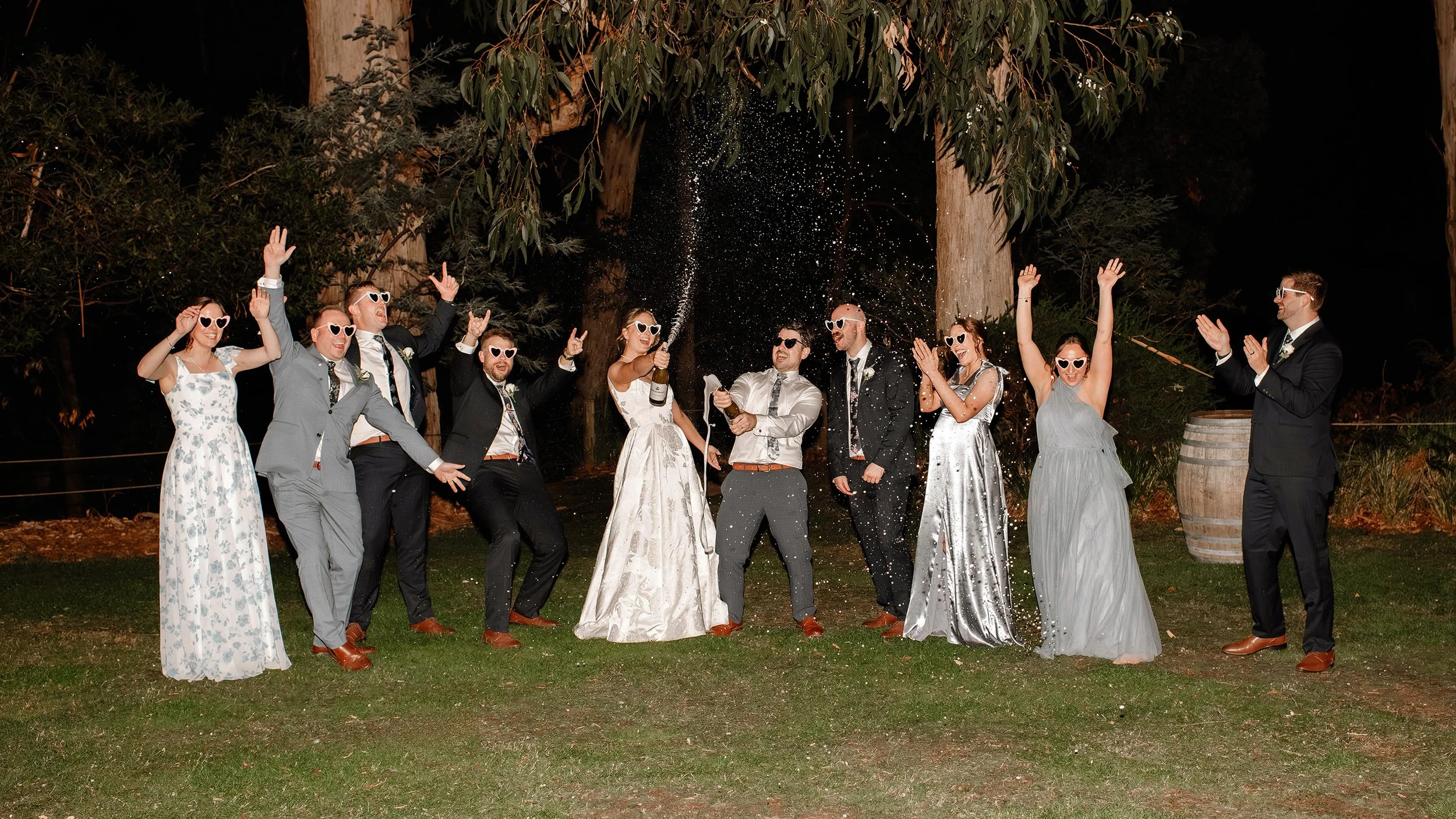 Wedding party in formal attire posing and celebrating on a lawn at night with hands raised in the air