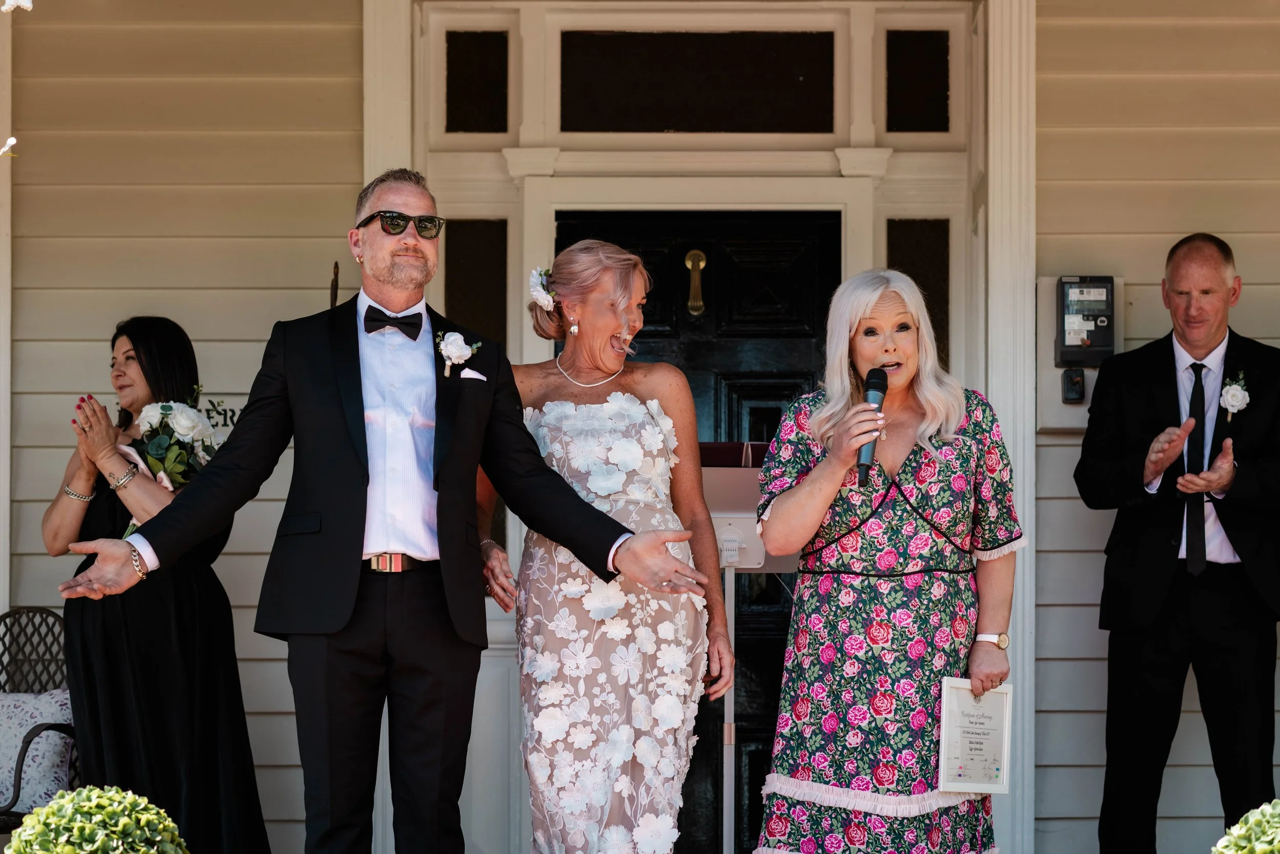 Bride and groom standing with a celebrant holding a microphone on the porch after their wedding ceremony
