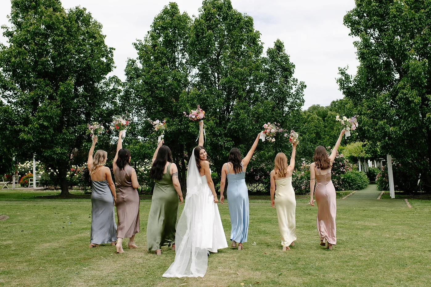 A bride and six bridesmaids walking outdoors on a grassy field, holding bouquets of flowers raised high. The bride wears a white wedding gown with a veil, and the bridesmaids wear colorful satin dresses. Tall trees and a cloudy sky are in the backgro