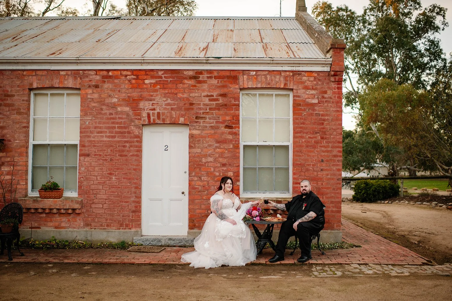 A newlywed couple sitting at a small round table outdoors in front of a red brick building, raising glasses for a toast. The bride wears a white wedding dress, and the groom wears all black. There are flowers on the table, and the building features t