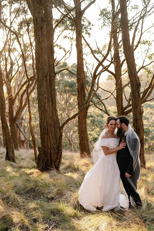 Bride in an off-the-shoulder wedding dress and groom in a dark suit embracing among tall trees in a sunlit forest