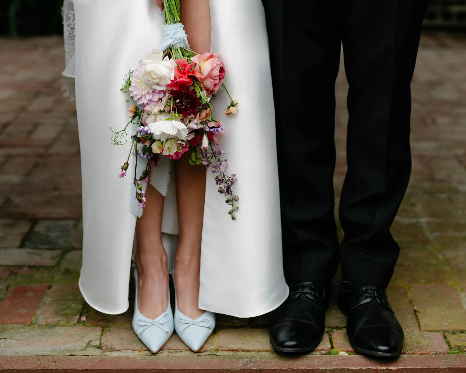 Close-up of a bride and groom's lower bodies, the bride holding a cascading bouquet of pink and white flowers, wearing white high heels, and the groom wearing black shoes, standing on a brick pathway. Daly Waters Mornington.