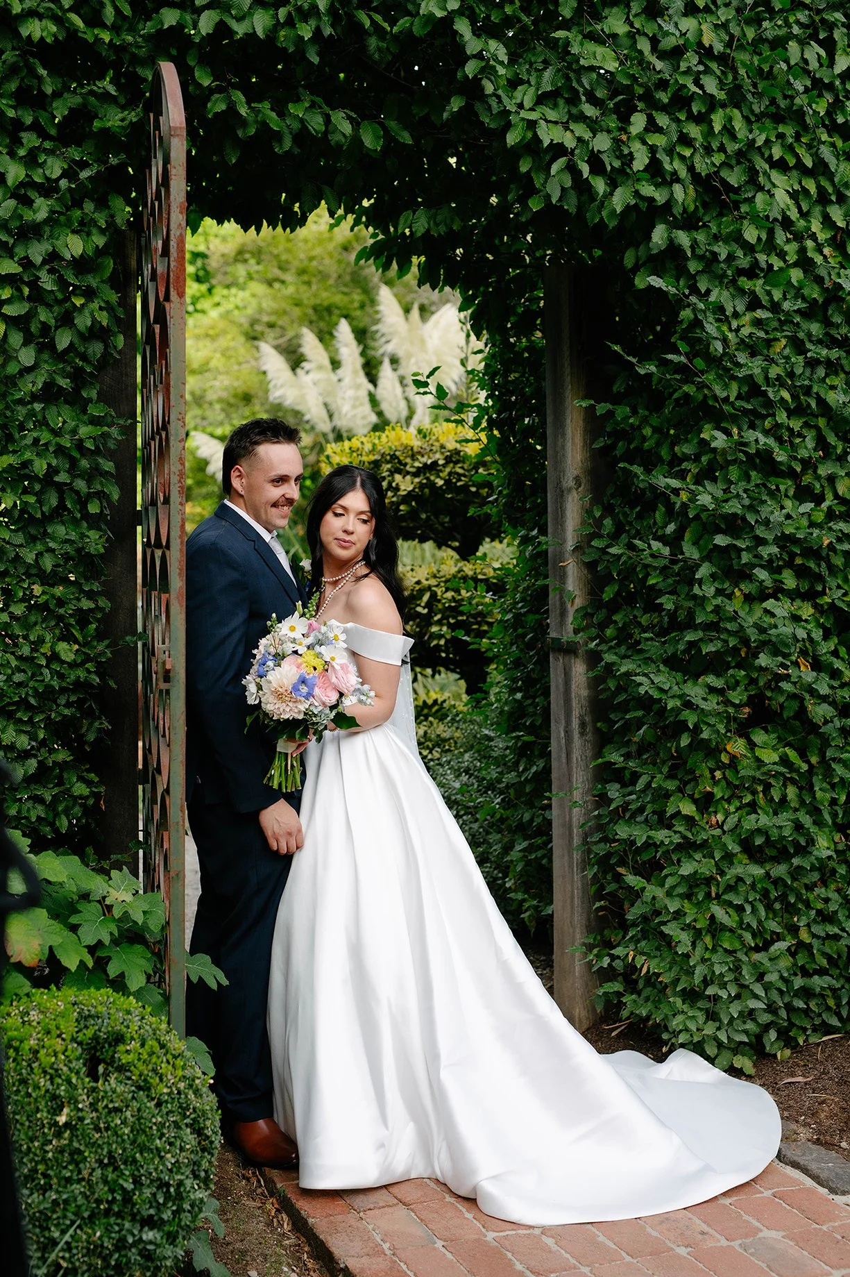 A bride and groom standing together in a garden archway, with the bride holding a bouquet of flowers, both dressed in wedding attire.
