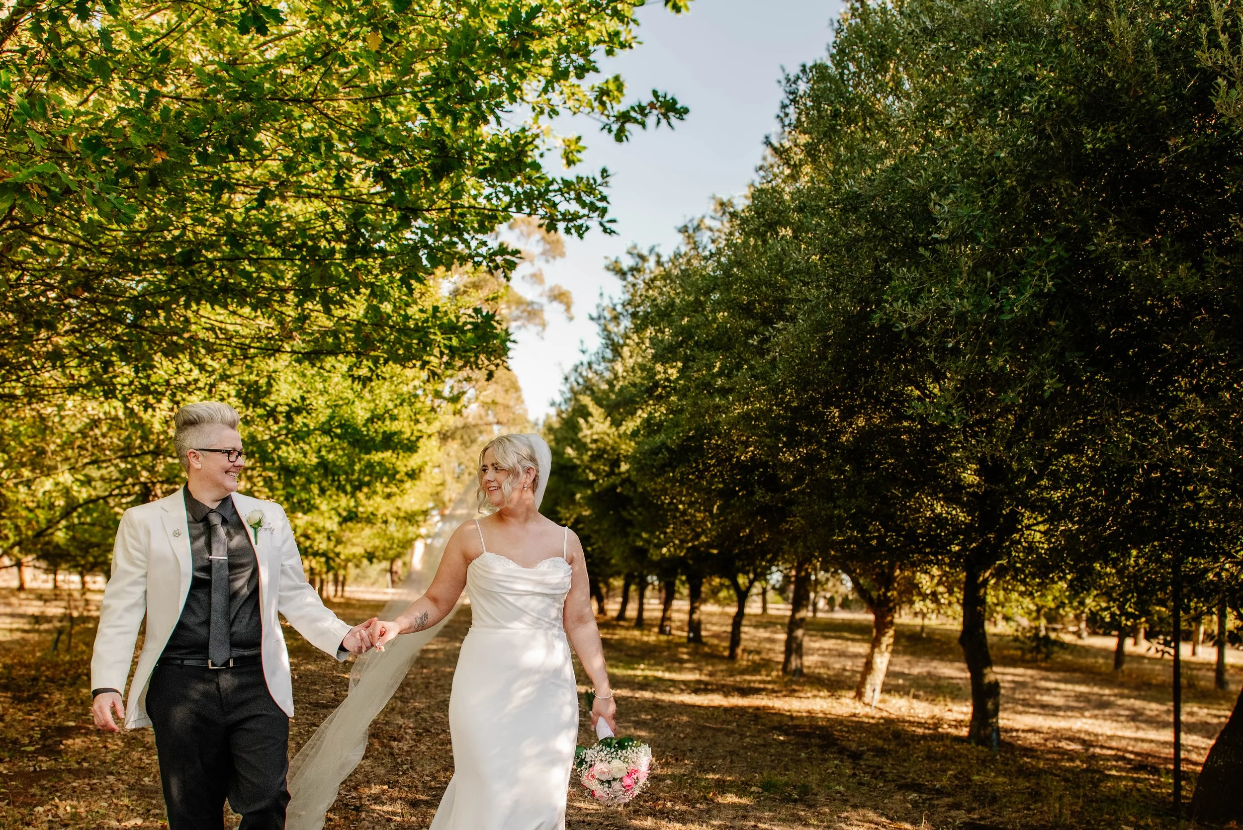Couple in wedding attire walking hand in hand through a sunny, green forest, smiling at each other with trees on both sides.